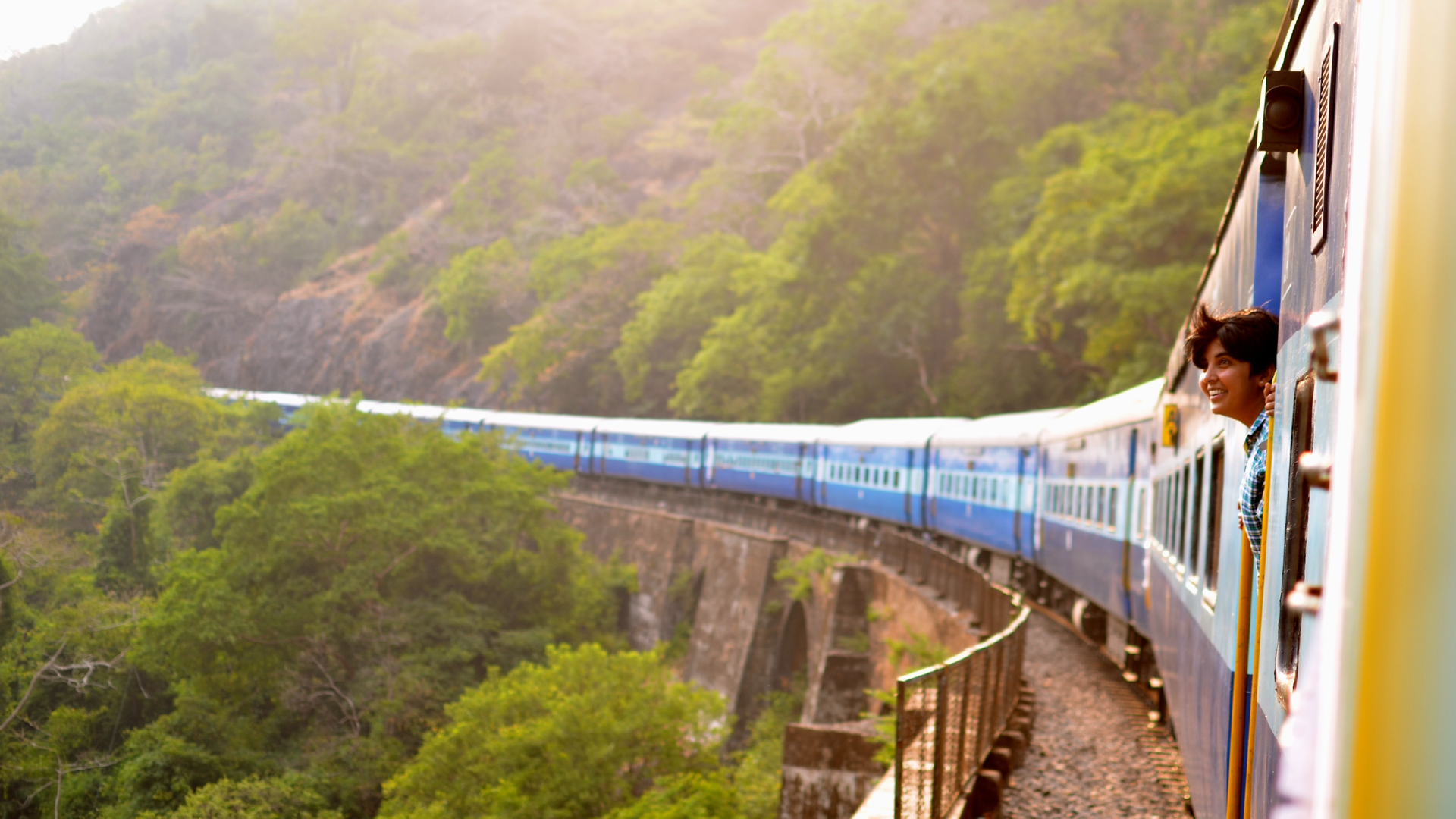 Zdjęcie przedstawia długi pociąg, który jedzie w scenerii górskiej. W oknie jednego z wagonów widoczna jest głowa kobiety.