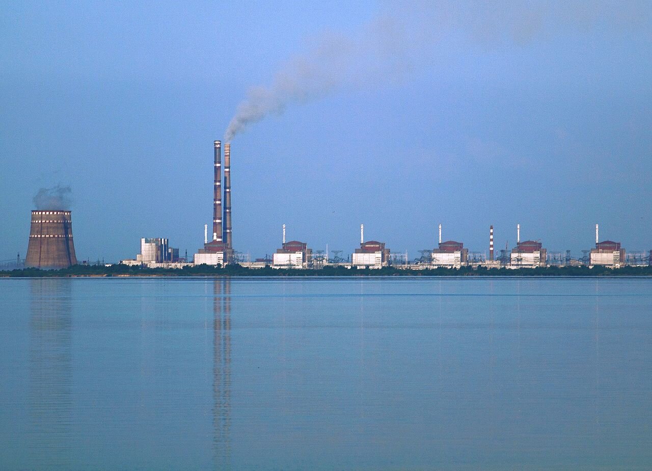 Zdjęcie przedstawia elektrownię jądrową, która umiejscowiona jest nad brzegiem zbiornika wodnego. Widoczne są szerokie, niskie kominy oraz wysokie i wąskie. Z kominów wydobywa się dym. Obok kominów liczne budynki w niskiej zabudowie.