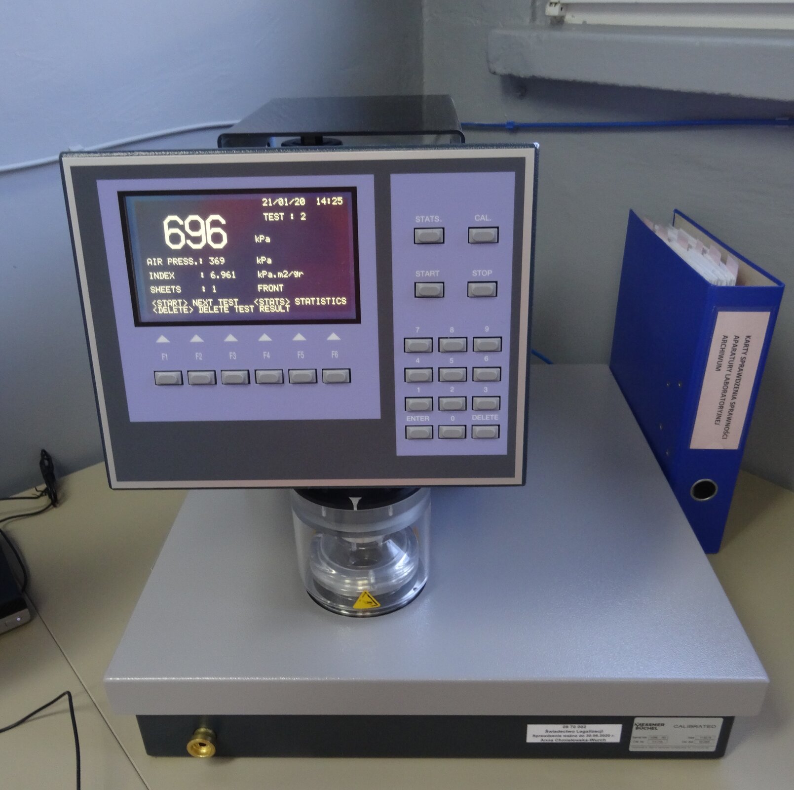 Zdjęcie przedstawia aparat do oznaczania odporności wytworów papierniczych na przepuklenie. Widoczne jest urządzenie z prostokątnym panelem sterowania, na którym znajduje się wyświetlacz z dokonanymi pomiarami oraz serią przycisków, w tym start, stop, enter, delate, przycisków numerycznych. Urządzenie ustawione jest na cylindrycznej, przeźroczystej nodze, która zamocowana jest prostopadłościennej podstawie. Całość stoi na blacie. Na prawo od urządzenia widoczny jest stojący segregator.