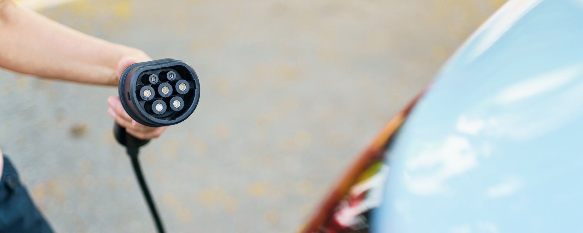 Zdjęcie przedstawia trzymaną w dłoni wtyczkę do zasilania elektrycznego samochodu. 