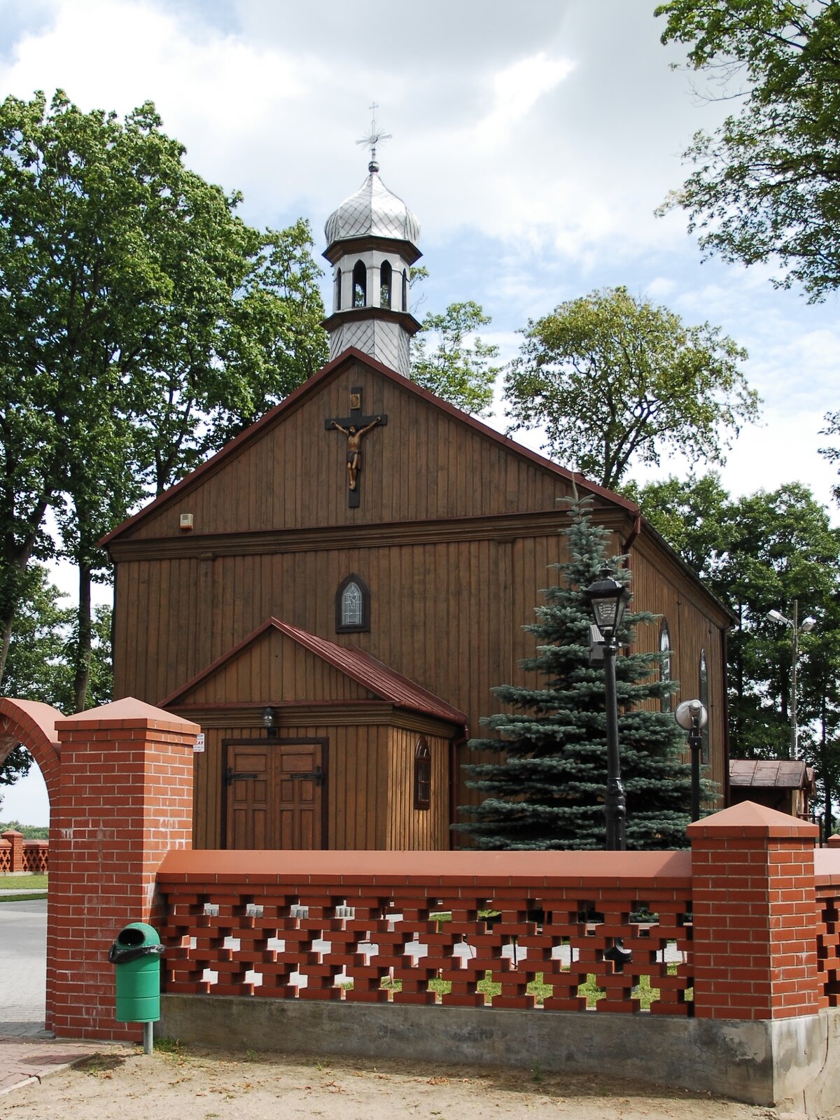 Zdjęcie przedstawia drewniany kościół, na czubku którego znajduje się srebrna wieżyczka z kopułą i krzyżem. Nad wejściem do kościoła wisi drewniany krzyż. Z prawej strony kościoła znajduje się zielona choinka. Kościół otoczony jest murowanym płotem, wykonanym z czerwonej cegły. Wokół kościoła znajdują się wysokie, zielone drzewa liściaste. Niebo jest błękitne, częściowo przysłonięte białymi chmurami. Zdjęcie zostało wykonane w świetle dziennym.