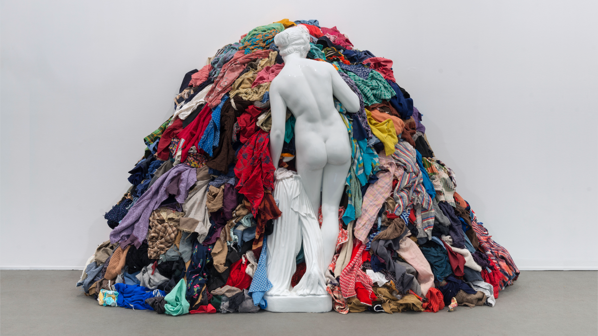 Zdjęcie przedstawia dzieło "Wenus wśród szmat". Jest to rzeźba przedstawiająca człowieka, stoi tyłem do kadru, jest naga. Rzeźba stoi przodem do ogromnej sterty ubrań. 