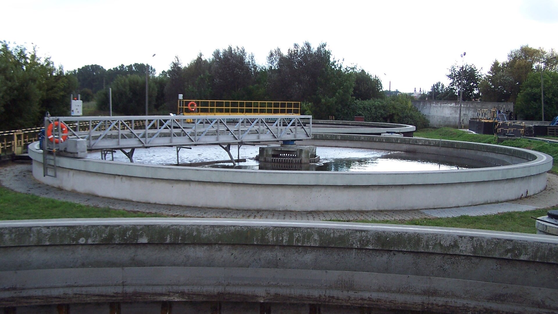 Fotografia przedstawiająca oczyszczalnię ścieków. Na pierwszym planie widoczny fragment zbiornika, za nim znajduje się okrągły zbiornik z cieczą, nad którym zbudowano mostek. Na dalszym planie jest jeszcze jeden okrągły zbiornik. W tle drzewa.