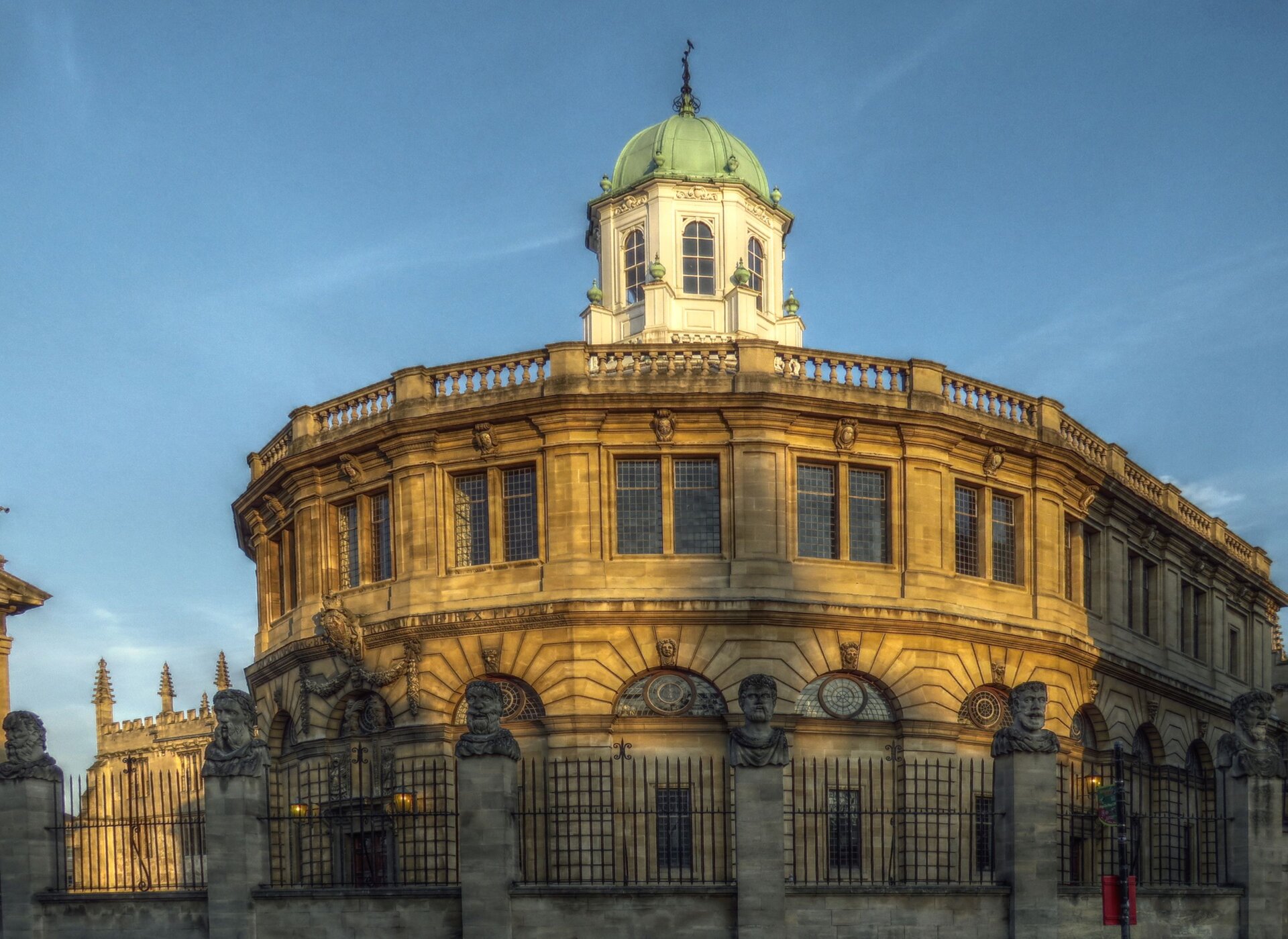 Ilustracja przedstawia budynek Teatru Sheldonian w Oksfordzie. Cały kadr wypełnia elewacja przednia, oparta na łuku oraz elewacja boczna. Budynek jest dwukondygnacyjny, z rytmicznie powtarzającymi się, rozmieszczonymi w tej samej odległości, otworami okiennymi. W dolnej kondygnacji mamy do czynienia z ciekawym rozwiązaniem: niewielkie prostokątne okna znajdują się wewnątrz łuków arkadowych oraz dodatkowe półokrągłe okna umieszczone są w górnej części tych samych łuków arkadowych. W górnej kondygnacji znajdują się podwojone, duże prostokątne okna, oddzielone pilastrami. Kondygnacje oddziela od siebie gzyms. Górna kondygnacja zwieńczona jest galerią, otaczająca niewidoczny z tej perspektywy, dach. Budynek ma niewielką, ośmioboczną kopułę pośrodku dachu, która ma półokrągło zakończone, duże okna ze wszystkich stron. Wokół budynku znajduje się metalowe ogrodzenie, z kamiennymi filarami zwieńczonymi popiersiami.