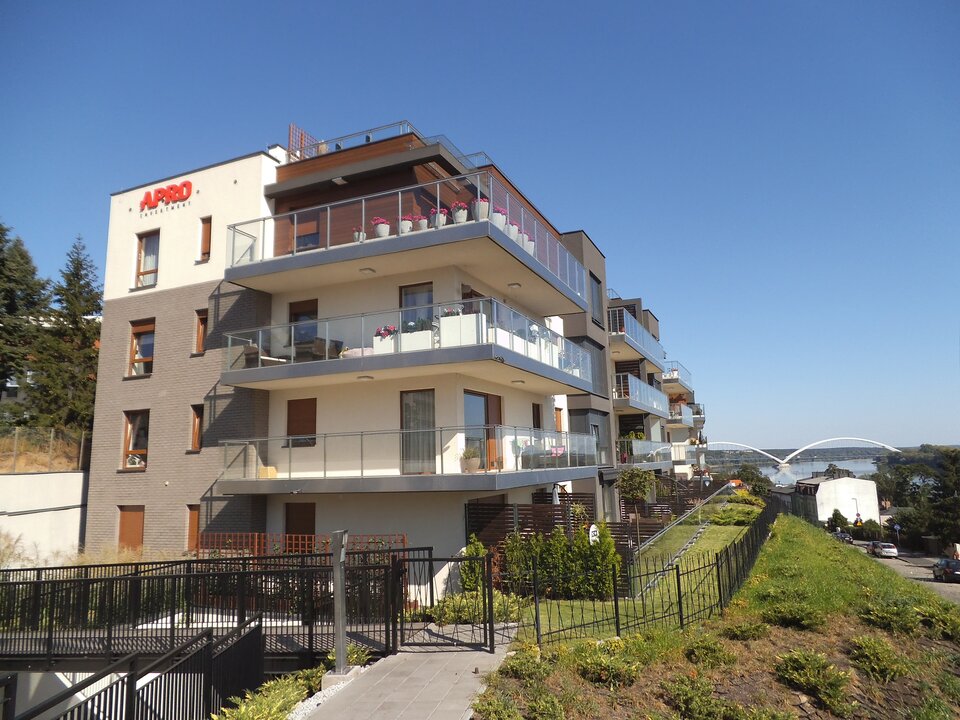 Zdjęcie przedstawia fragment zabudowy wielorodzinnej przy ulicy Lubickiej w Toruniu. Bloki są umieszczone bokiem do obiektywu aparatu, są kształtem zbliżone do sześcianu. Posiadają parter i trzy kondygnacje. Z górnych kondygnacji wychodzi się na przeszklony balkon, z mieszkań na dolnej kondygnacji jest wyjście na patio. Osiedle ogrodzone jest niskim, czarnym płotem. 