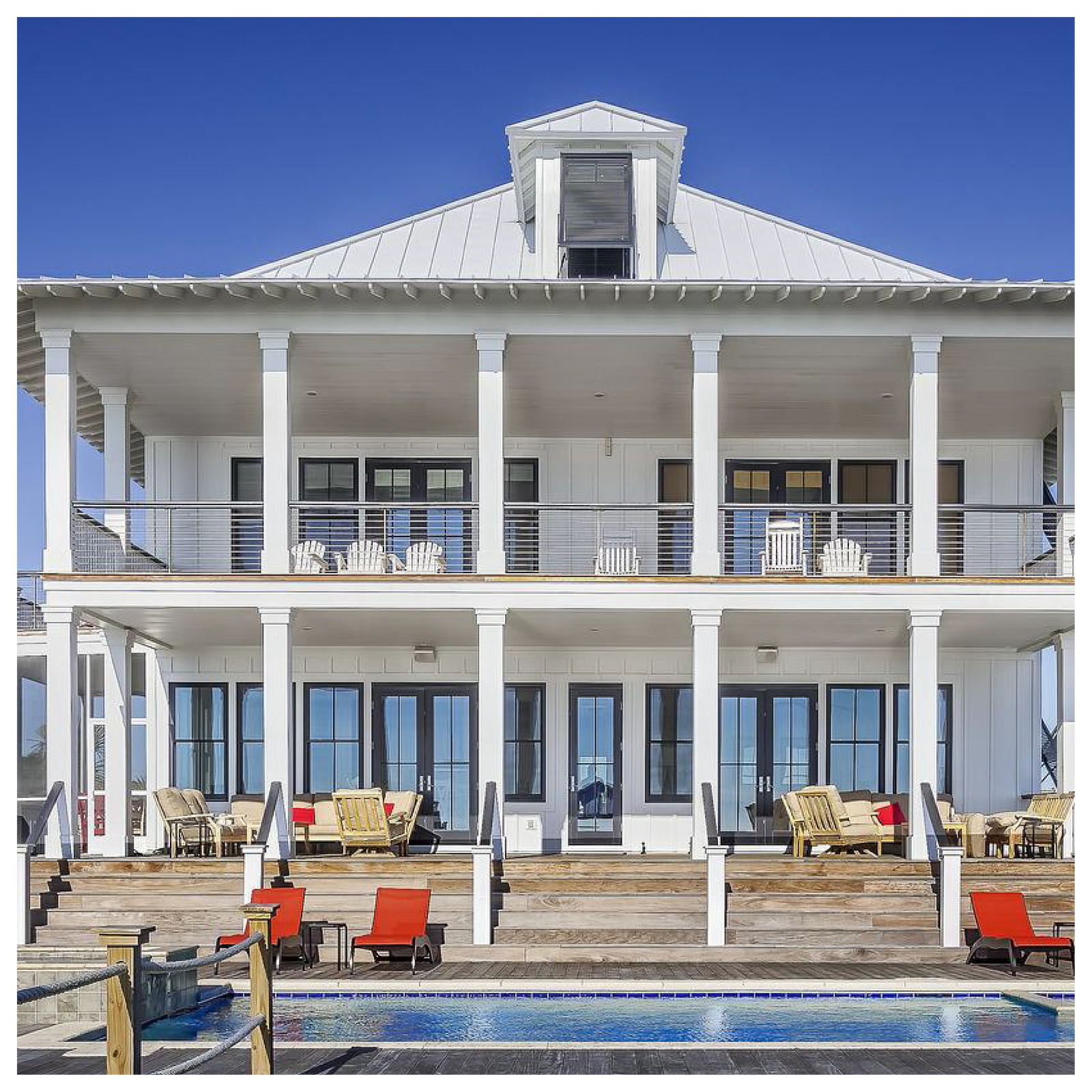 Na zdjęciu widoczny jest duży, biały, dwupiętrowy dom z tarasem i basenem. Na pierwszym piętrze jest taras z krzesłami. Na parterze jest również taras z krzesłami oraz schody prowadzące do basenu. Nad basenem są czerwone leżaki.