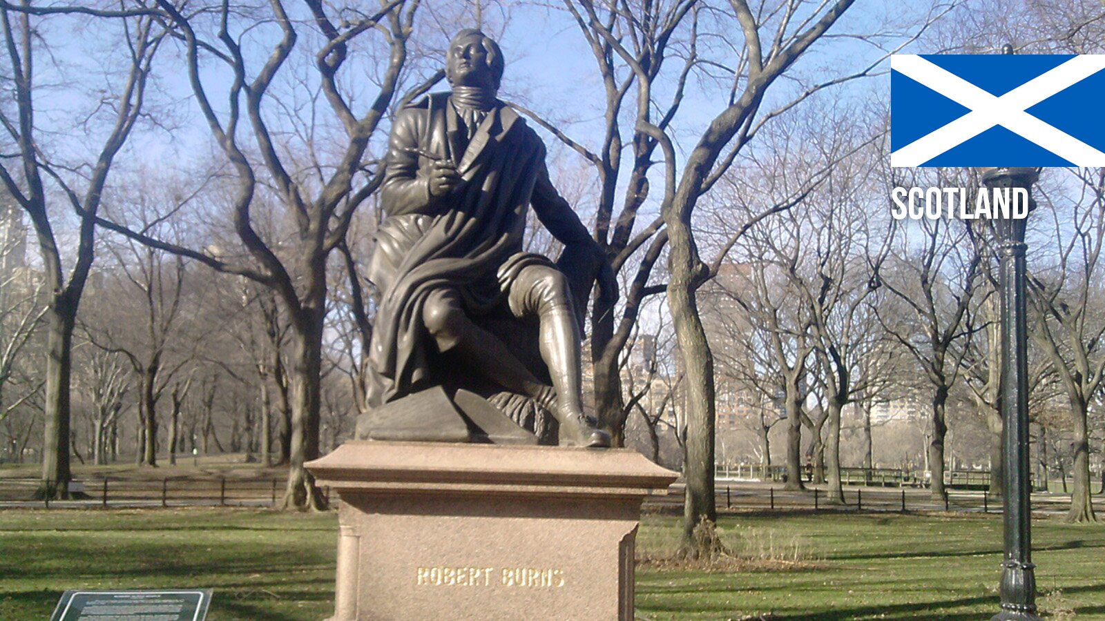 Zdjęcie przedstawia pomnik siedzącego mężczyzny na kamiennym postumencie. Pomnik stoi w parku. Pomnik przedstawia mężczyznę w płaszczu, siedzącego na podwyższeniu, z głową skierowaną do góry, jakby patrzył w niebo. Jego lewa ręka jest lekko ugięta. Na kamiennym postumencie widoczne jest imię i nazwisko: Robert Burns. W prawym górnym rogu  widoczna jest szkocka flaga. 