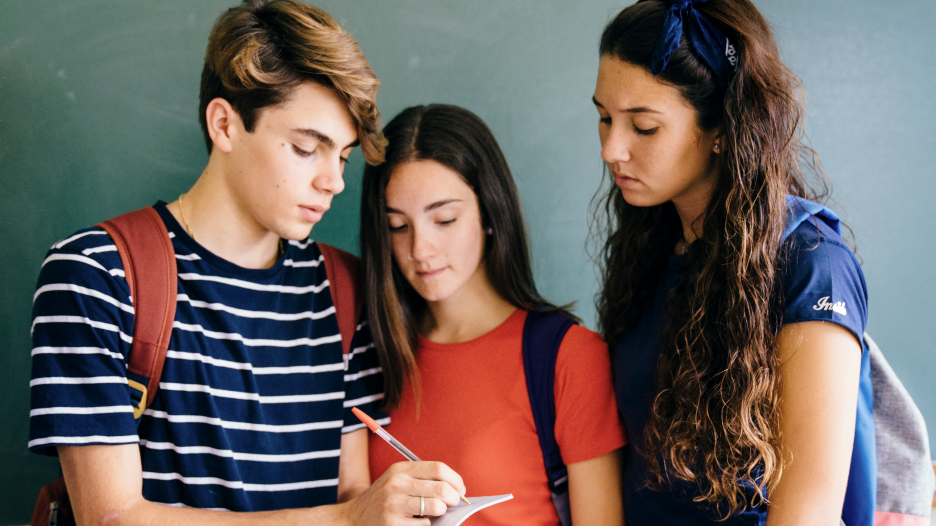 Zdjęcie przedstawia troje nastolatków. Po lewej znajduje się chłopak w koszulce w paski i z plecakiem na plecach. Chłopak trzyma w ręku długopis i notuje coś na kartce. Pokazuje tę kartkę dziewczynom stojącym obok niego. Dziewczyna w środku na długie włosy i jest najniższa z całej trójki. Dziewczyna przygląda się na to, co chłopak zapisuje na kartce. Dziewczyna po prawej jest najwyższa, ma lekko kręcone włosy i również spogląda na to, co zapisuje chłopak na kartce.