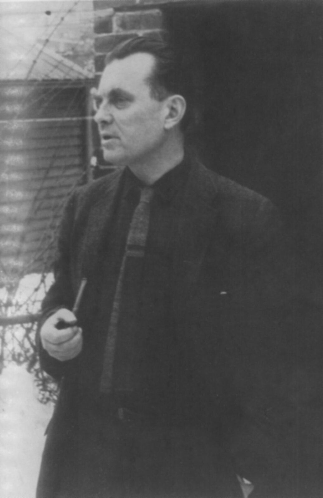 Zdjęcie przedstawia mężczyznę w średnim wieku. Mężczyzna stoi, trzyma w prawej dłoni fajkę. Jest ubrany w garnitur.