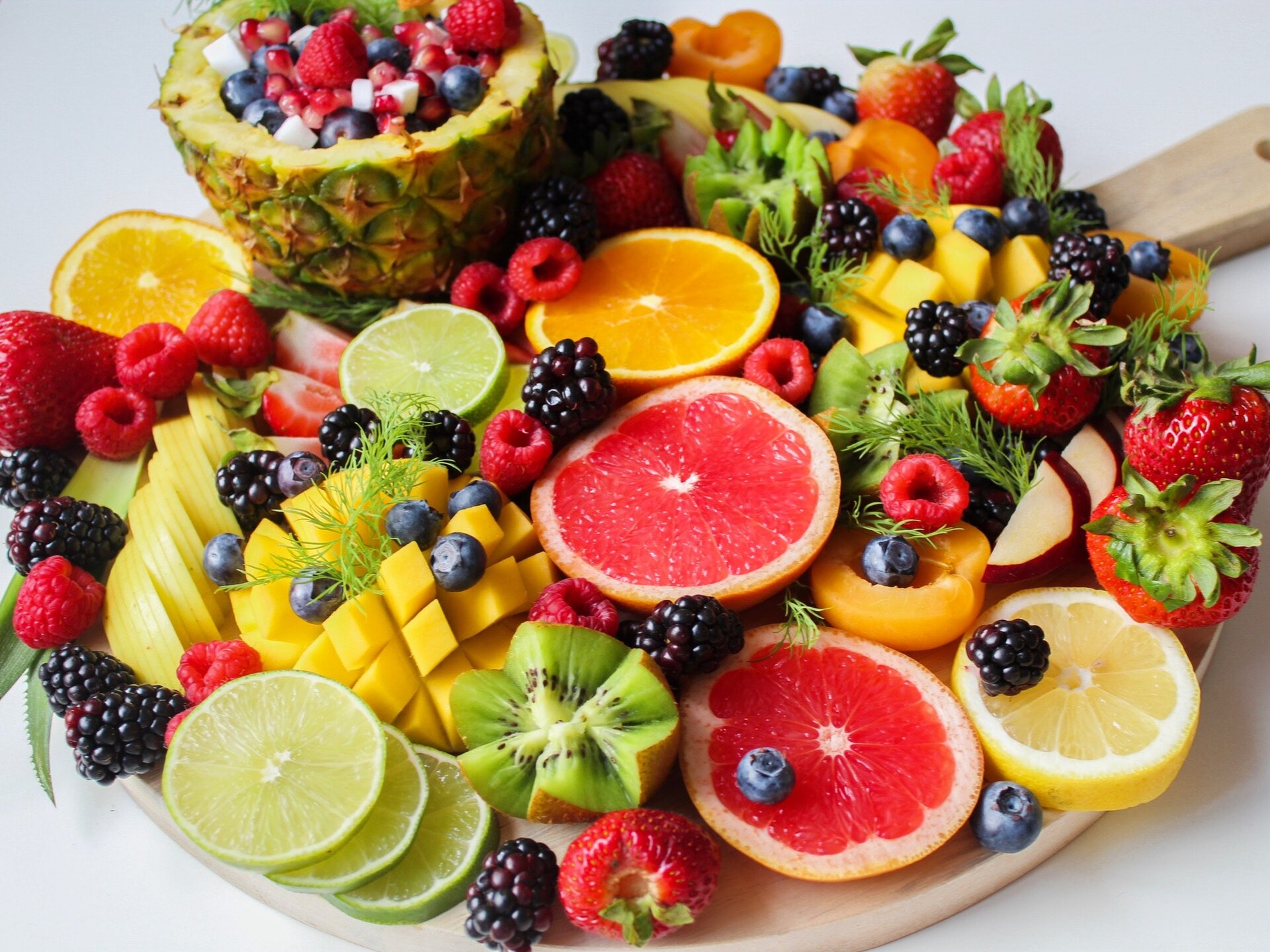 Zdjęcie [przedstawia deskę z wieloma owocami: pomarańczami, grejpfrutami, kiwi, brzoskwiniami, mango, malinami, jeżynami, ananasem, cytrynami.