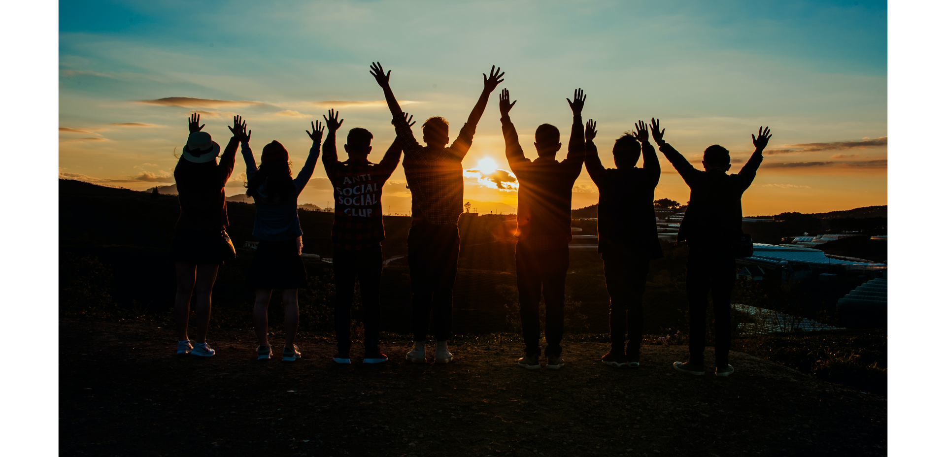 Zdjęcie przedstawia ciemne sylwetki siedmiorga ludzi. Wszyscy trzymają ręce uniesione nad głową. Zdjęcie zostało wykonane pod słońce. Słońce chyli się ku zachodowi.