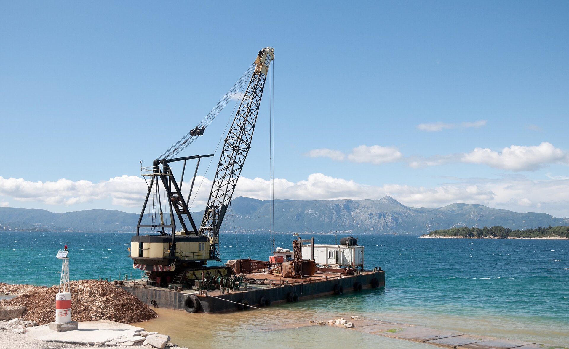 Na zdjęciu widoczna zatoka morska. Przy brzegu platforma do rozładunku. Na platformie dźwig z wysuniętym do góry ramieniem. Na szczycie zaczepiony jest system lin, które unoszą ładunek.