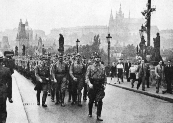 Zdjęcie przedstawia marsz w Pradze. Kolumna mężczyzn idzie środkiem mostu. Przewodzi im mężczyzna w mundurze z odznaczeniami, w czapce wojskowej. Po obu stronach mostu, po chodnikach, idą ludzie. Przy chodnikach są latarnie uliczne. Z prawej strony stoi drewniany krzyż z figurą Jezusa. W tle znajdują się zabudowania miasta.