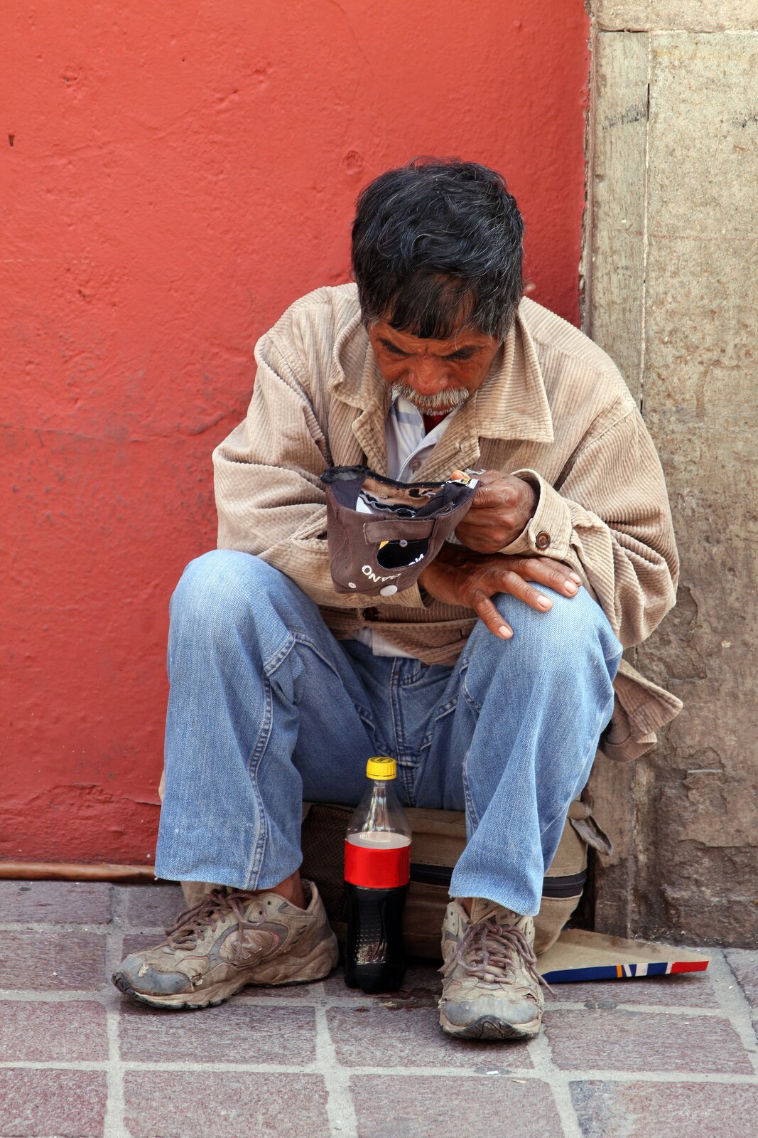 Kliknij, aby powiększyć. Zdjęcie przedstawia starszego mężczyznę siedzącego pod ścianą i proszącego o datki. Ubrany jest w beżową, sztruksową kurtkę, białą koszulę, niebieskie dżinsowe spodnie i mocno zniszczone, jasne, sportowe buty. Ma ciemne włosy i siwe wąsy oraz brodę. Siedzi na beżowej walizce, która leży na bruku. Ręce ma oparte o kolana. W lewej dłoni trzyma brązową czapkę z daszkiem, odwróconą tak, aby wrzucać do niej datki. Przed nim stoi butelka z napojem. Mężczyzna plecami oparty jest o ścianę, po lewej stronie czerwoną, po prawej w kolorze piaskowca.