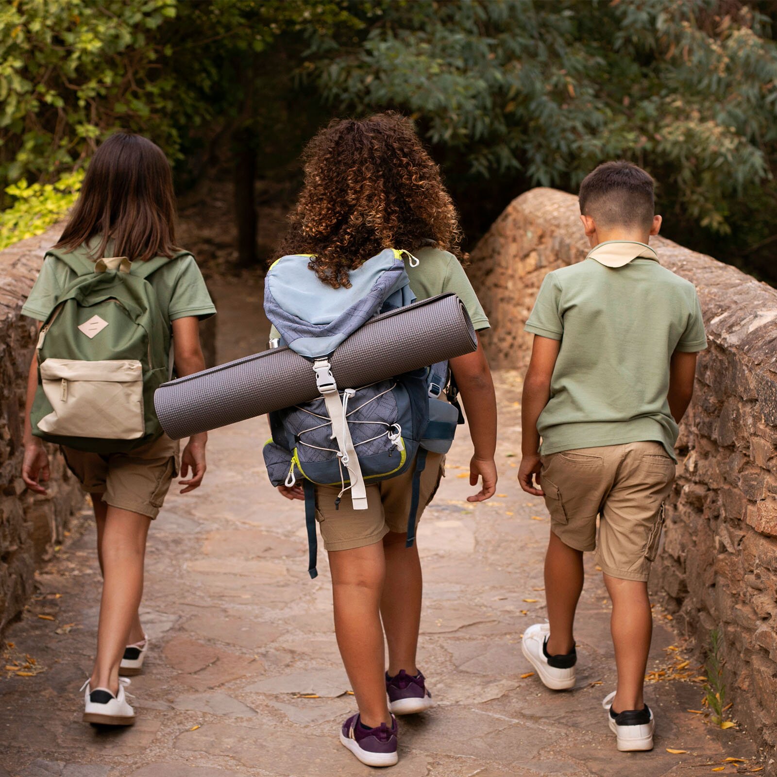 Zdjęcie przedstawia trójkę dzieci, w ujęciu od tyłu, idących wąskim kamiennym mostkiem. Wszyscy ubrani są w beżowe spodenki, bladozielone koszulki z krótkim rękawem i sportowe buty. Od lewej strony widoczna jest dziewczynka z długimi, ciemnymi włosami, z zielono‑kremowym plecakiem na plecach. W środku widoczna jest dziewczynka z kręconymi, ciemnymi włosami, z plecakiem na plecach, przez klapę którego przełożona jest szara karimata. Po prawej stronie widoczny jest chłopiec z krótkimi, ciemnymi włosami. W tle widoczne są drzewa.