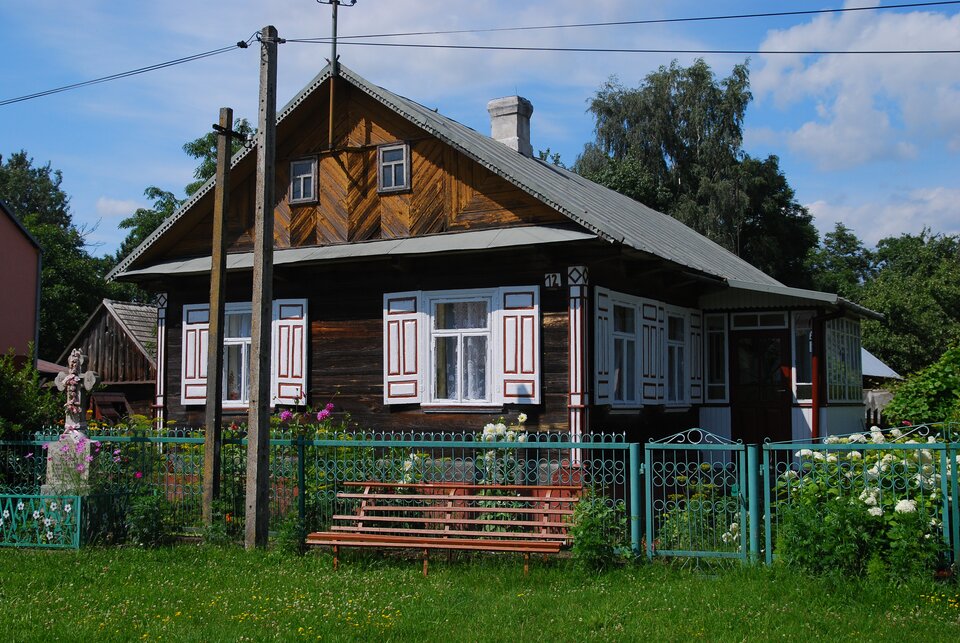 Zdjęcie przedstawia niewielki dom z okiennicami i spadzistym dachem. Dom jest drewniany. Dookoła budynku jest zielony płot. Przed ogrodzeniem stoi kapliczka i ławka. Przed domem i obok budynku rosną kwiaty, za domem drzewa.  