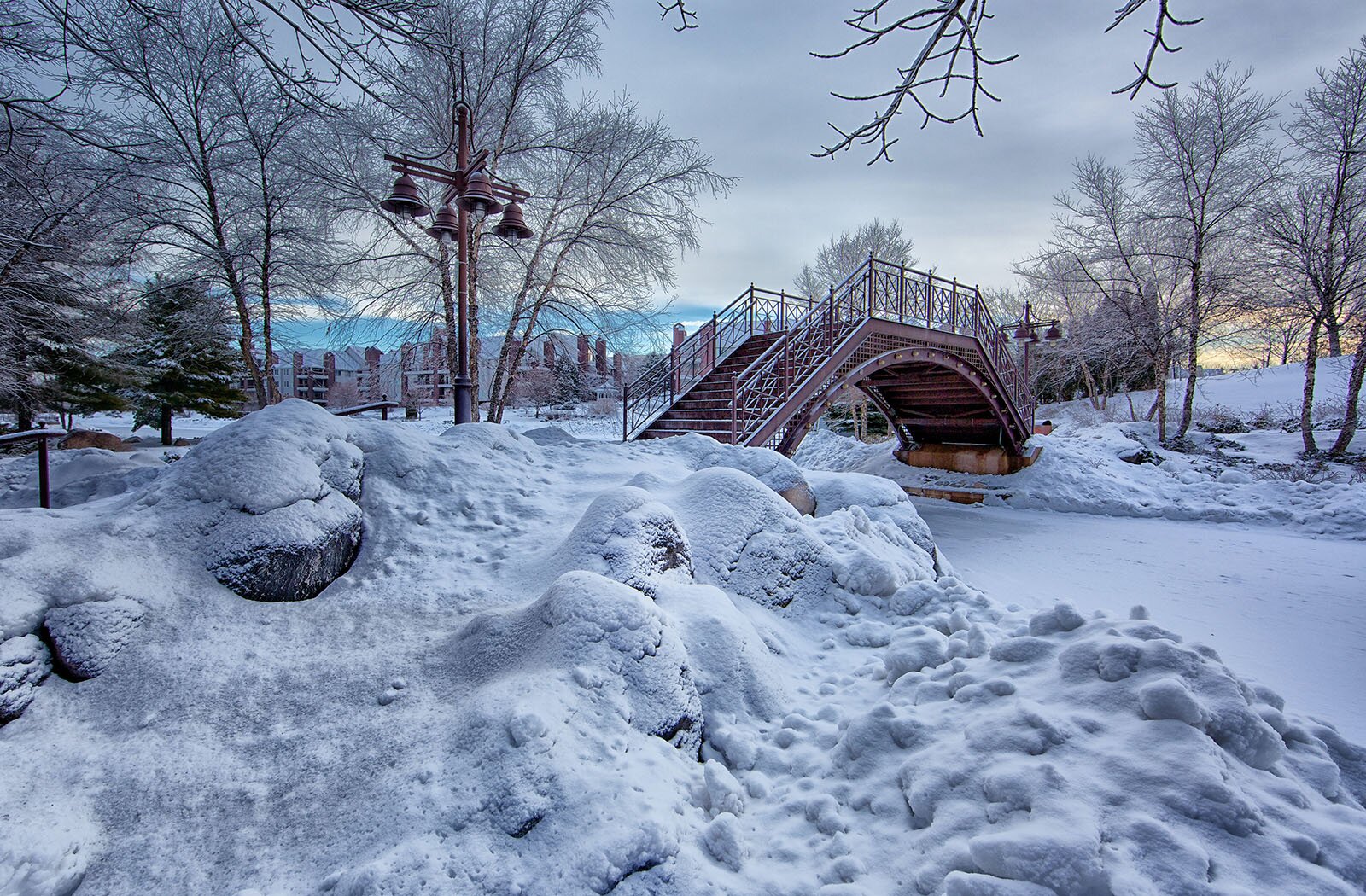 Zdjęcie przestawia okolicę zasypaną śniegiem. Widoczny jest stalowy, wąski i wysoki mostek nad zamarzniętą rzeką. Do mostka prowadzą schody, przed którymi z obu stron widoczne są latarnie uliczne, każda z czterema lampami w kształcie dzwonów. Wokół widoczne są zaspy śniegu, bezlistne, oszronione drzewa oraz ośnieżone drzewa iglaste.  W tle widoczne są szaro‑białe chmury, a tuż nad horyzontem skrawek niebieskiego nieba.