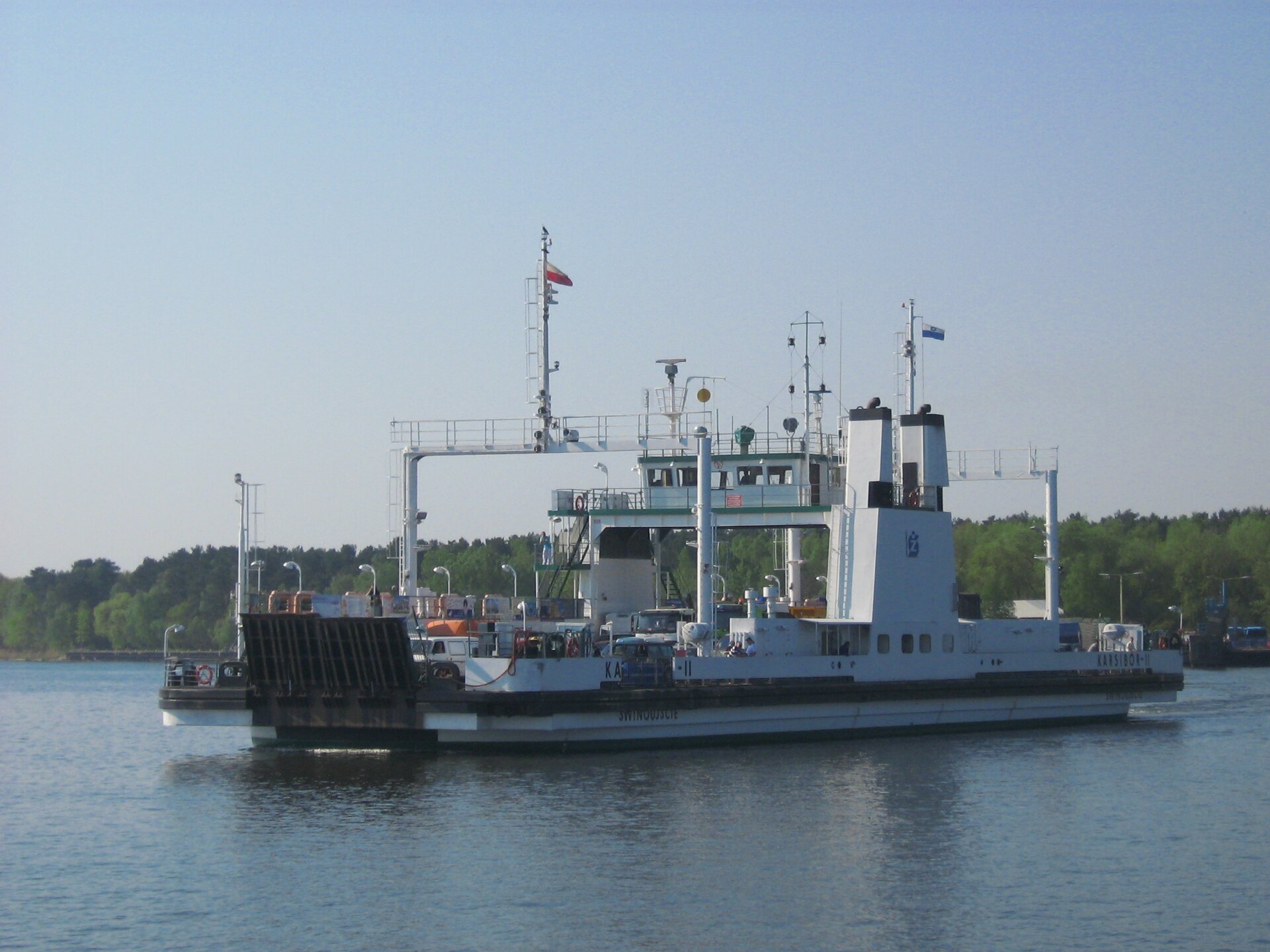Zdjęcie przedstawia płynący prom. Na jego froncie widnieje zamontowana, opuszczana rampa wjazdowa. Na jego pokładzie kilka zaparkowanych pojazdów. W tle las oraz elementy infrastruktury portowej, na której powiewa flaga Polski.