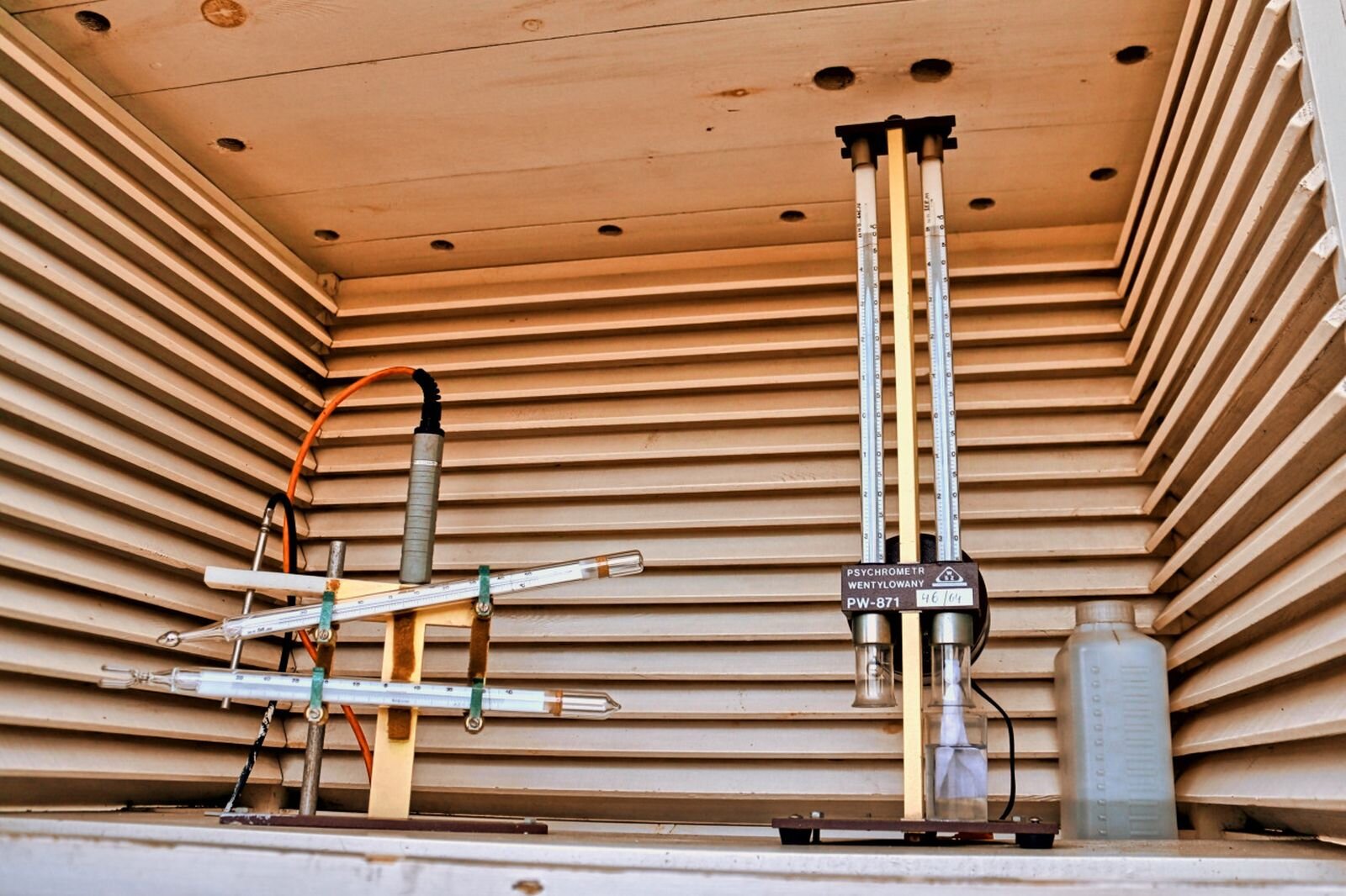Zdjęcie przedstawia wnętrze klatki meteorologicznej. Po lewej znajdują się termometry: maksymalny i minimalny, a po prawej – rodzaj higrometru, przyrządu do pomiaru wilgotności powietrza. Termometry umiejscowiony jest w poziomie, a higrometr w pionie.
