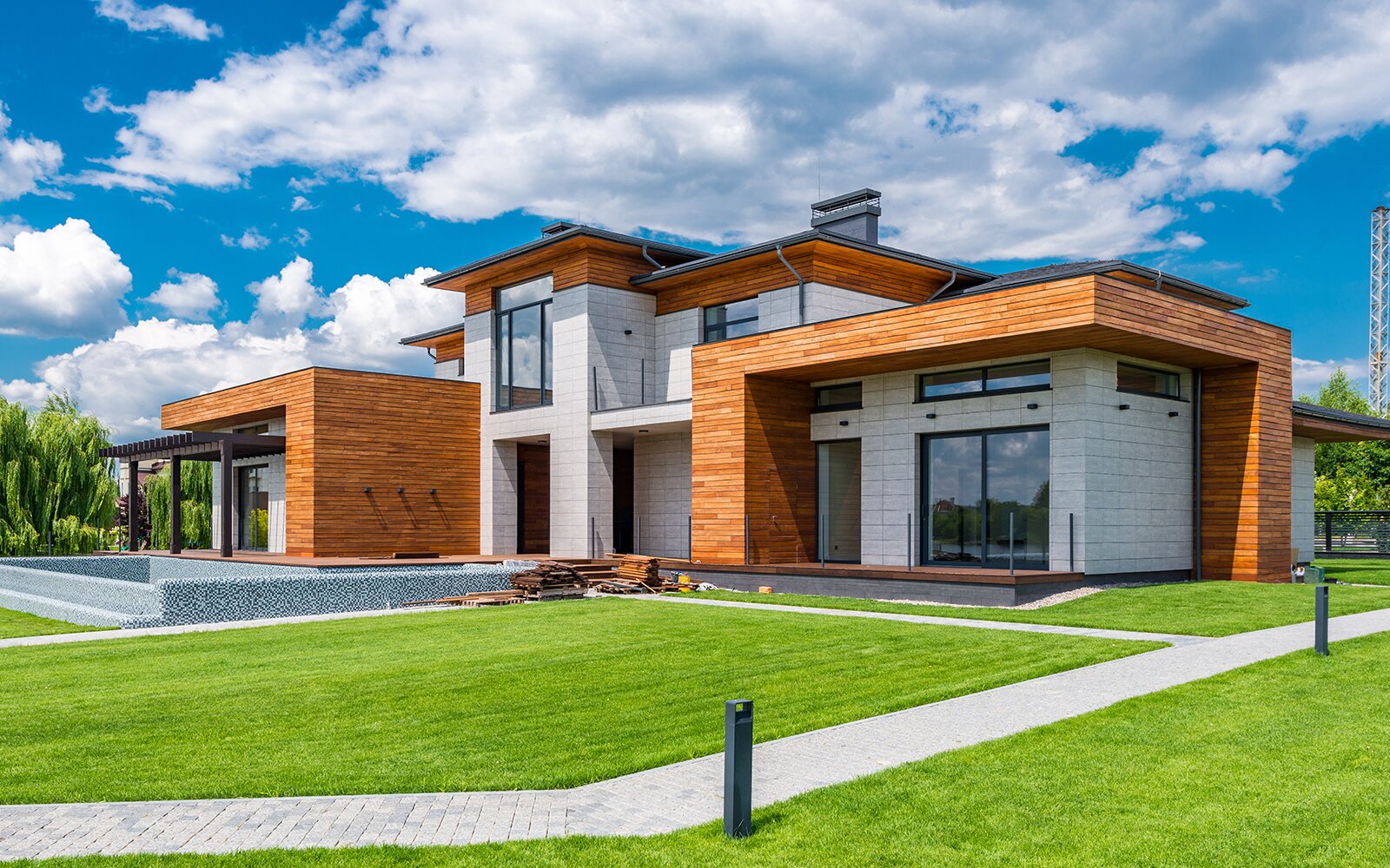 Zdjęcie przedstawia nowoczesny dom zbudowany z białej cegły i drewnianych paneli z płaskim dachem. Dom składa się z trzech części. Ma kilka dużych okien. Transkrypcja nagrań znajduje sie poniżej. 