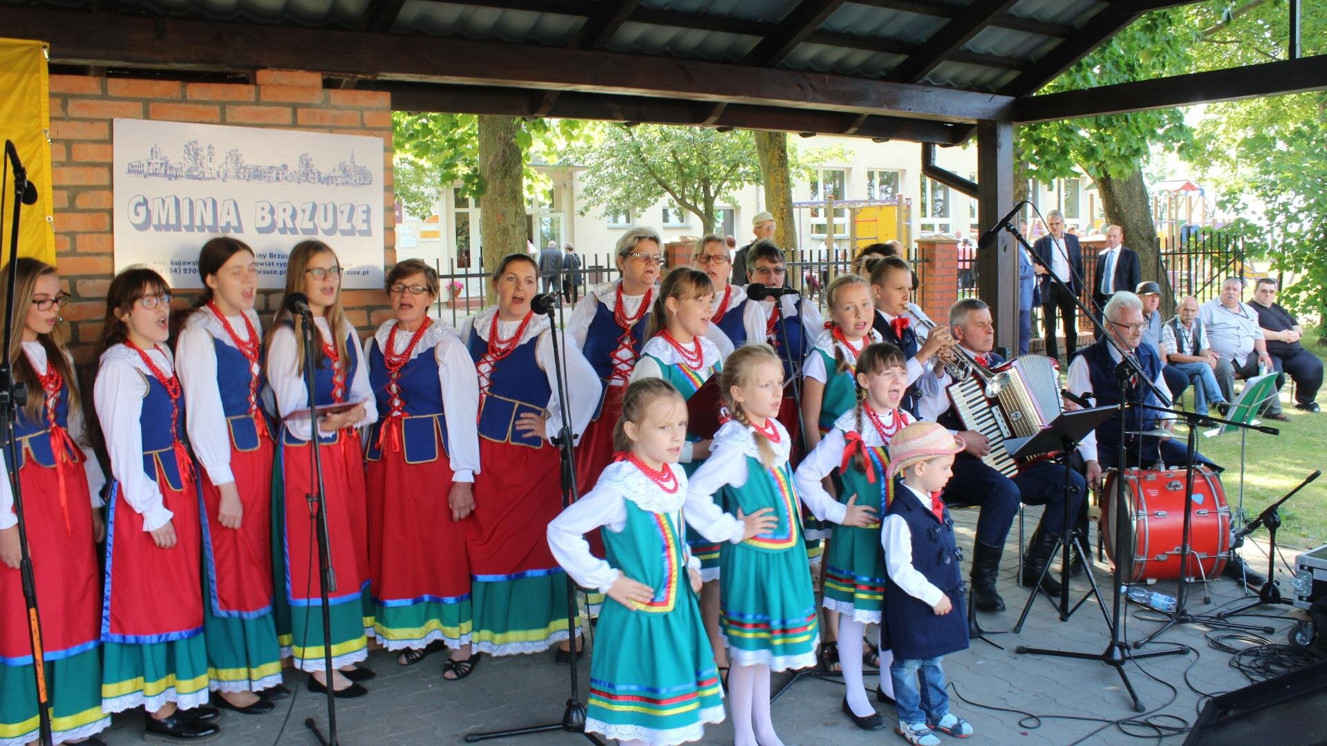 Zdjęcie przedstawia zadaszoną scenę, na której stoją kobiety ubrane w stroje ludowe oraz mężczyźni grający na instrumentach. Kobiety znajdują się z lewej strony zdjęcia i ubrane są tak samo: czerwono‑zielona spódnica, biała koszula, niebieska kamizelka, czerwone korale. Kobiety mają otwarte usta. Przed nimi stoją młode dziewczyny, które ubrane są w zielone spódniczki, białe koszule, zielone kamizelki i czerwone korale. Przed nimi stoi mały chłopiec, który ma na sobie niebieskie spodnie, białą koszulę, granatową długą kamizelkę i słomkowy kapelusz. Z prawej strony zdjęcia widoczni są siedzący mężczyźni, z których jeden z nich gra na akordeonie, a drugi trzyma między nogami bęben. W tle za nimi widoczne są pojedyncze osoby ubrane w codzienne stroje. Za kobietami widoczna jest ceglana ściana, na której przypięty został plakat z napisem "GMINA BRZUZE". Zdjęcie zostało wykonane w świetle dziennym.