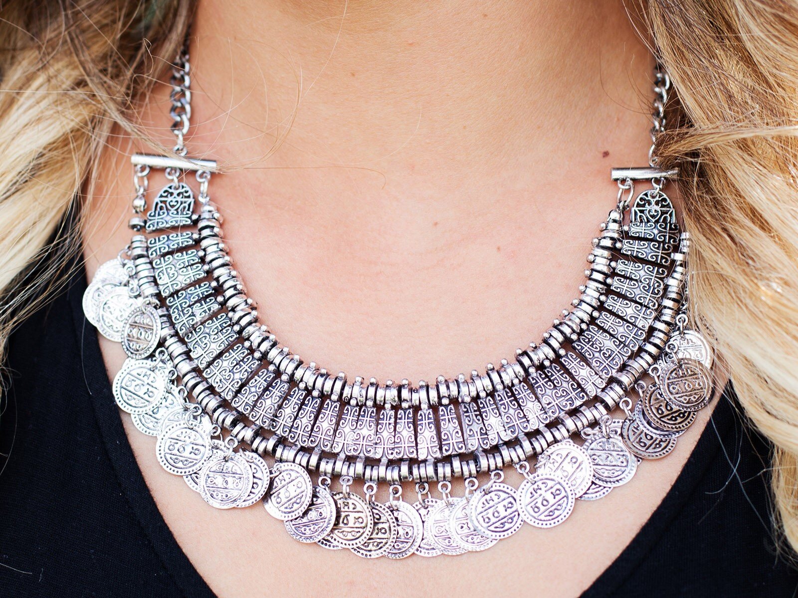 Zdjęcie przedstawia kobiecy dekolt, na którym widoczny jest srebrny naszyjnik w stylu orientalnym, złożony z kilku dekoracyjnych elementów i zamocowany na srebrnym łańcuszku. Na górze znajduje się szeroki pas z pierścieniami i ozdobnymi rombami, a poniżej zawieszone na nim są okrągłe wisiorki w kształcie monet.