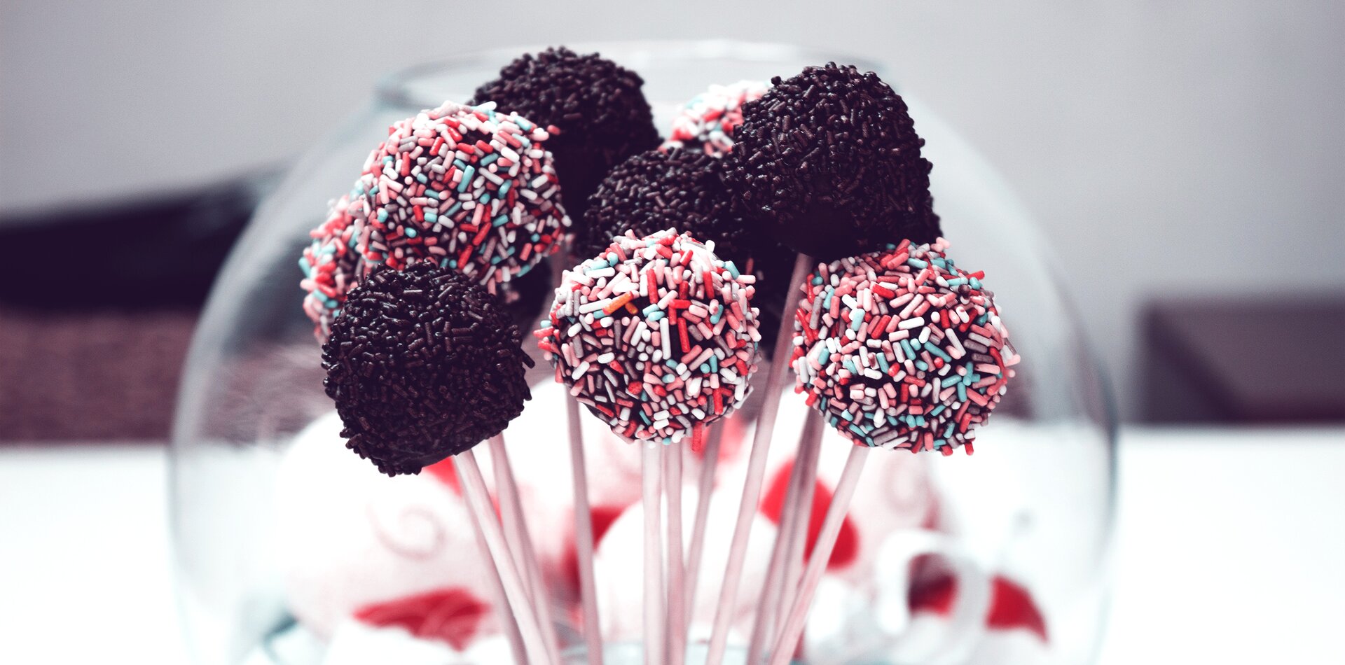 Zdjęcie kilku lizaków w kształcie kolorowych kulek na patykach. Połowa lizaków ma posypkę czekoladową, a połowa wielobarwną. Na drugim planie widać inne słodycze w szklanym naczyniu o kształcie kuli. 