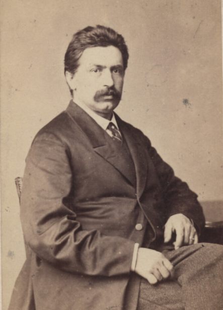 Ilustracja przedstawia mężczyznę w średnim wieku siedzącego prawym pólprofilem do widza. Ciemne włosy zaczesane do tyłu, sumiaste wąsy. Ubrany jest w garnitur, kamizelkę, koszule i krawat. Mężczyzna siedzi na krześle. lewa ręka oparta o blat stołu.