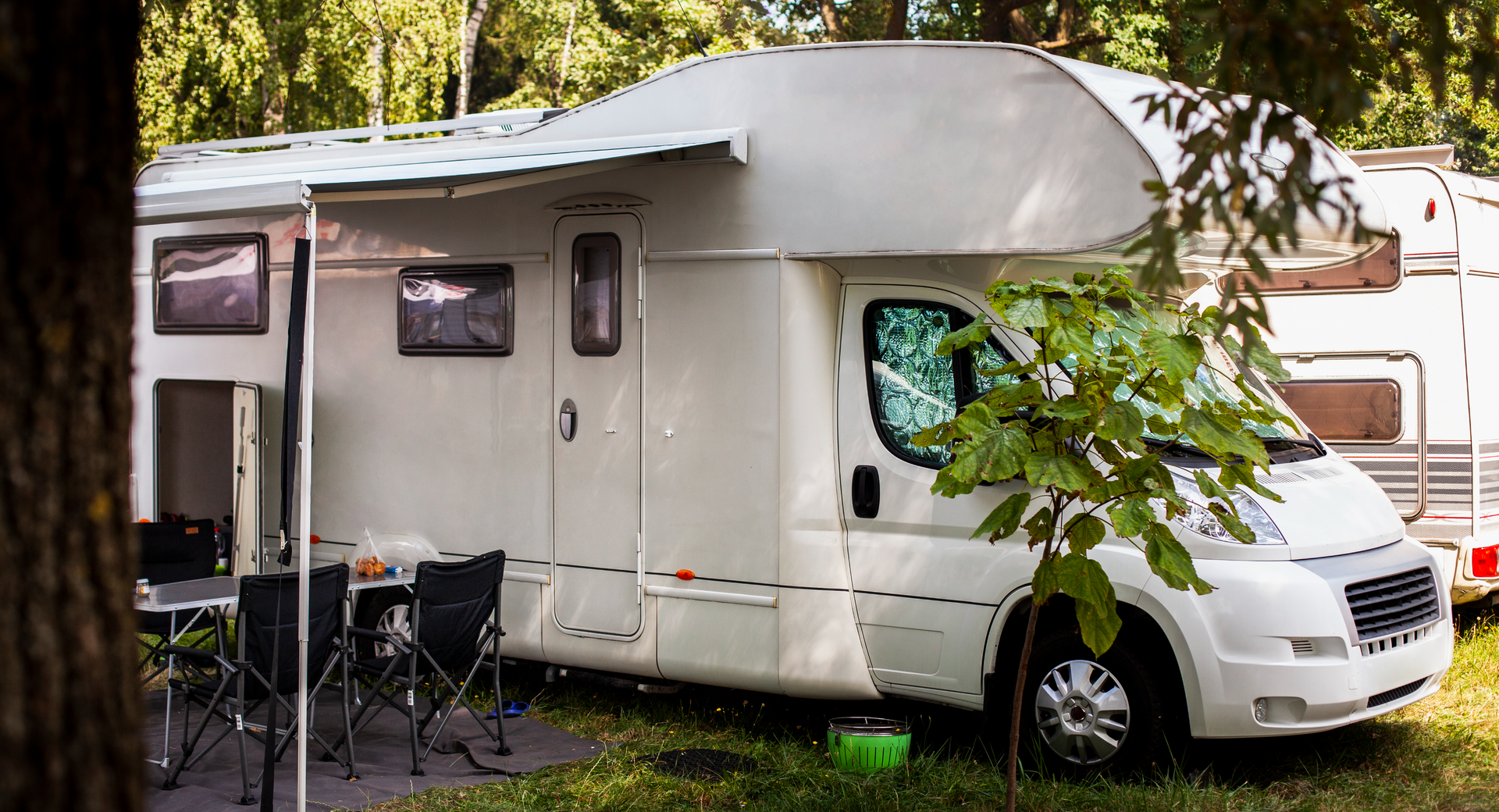 Na zdjęciu widoczny jest zaparkowany kamper z markizą, pod którą stoi rozkładany stół i krzesła.