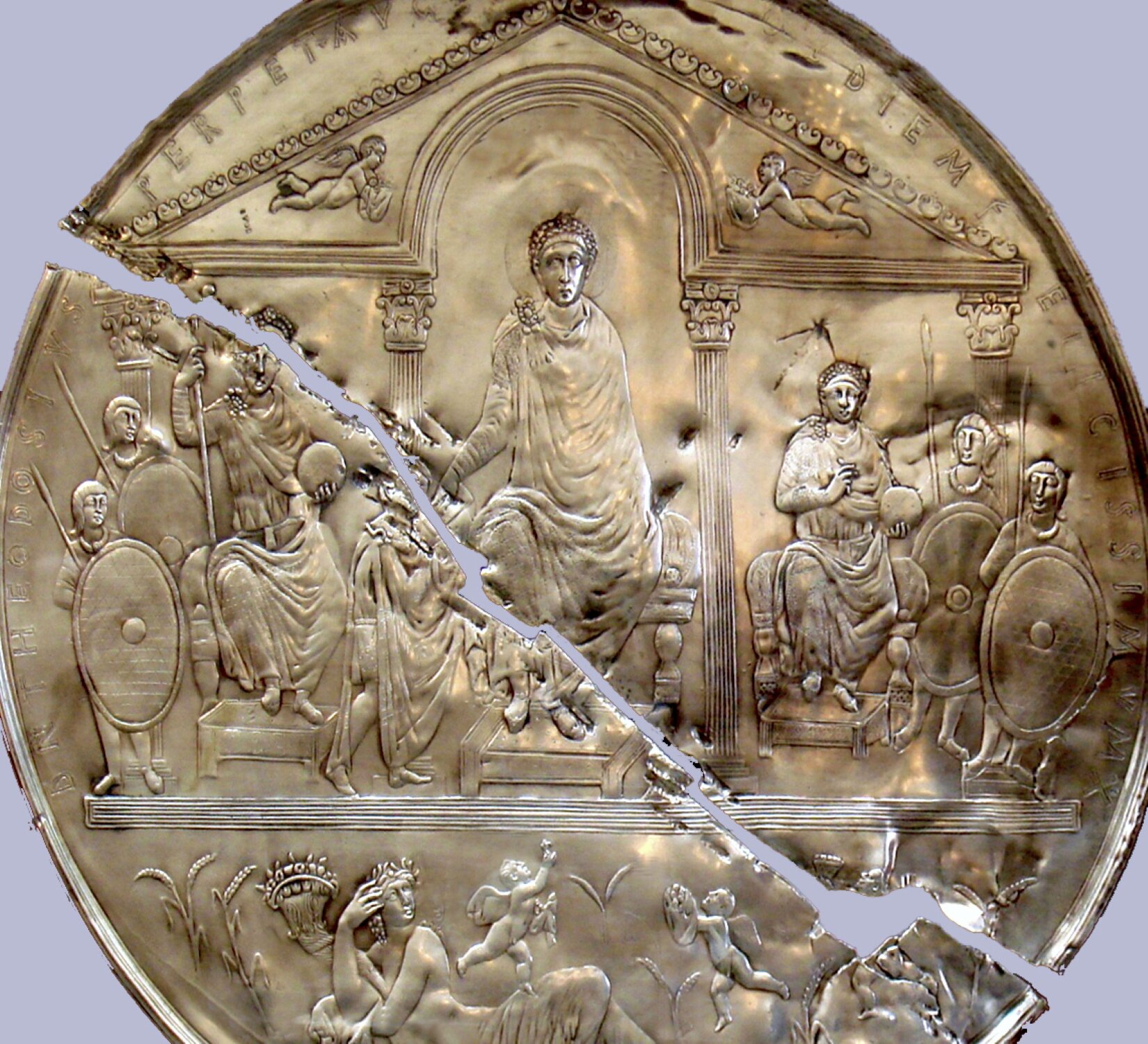 Fotografia przedstawia okrągły, złoty przedmiot przypominający pęknięty wpół dysk, ukazany na tle szarej ściany. Na jego powierzchni wykonano zdobienia przedstawiające postacie w budynku z łukowym sklepieniem podtrzymanym kolumnami. Tympanon ozdobiono wizerunkami postaci – nagich dzieci ze skrzydłami na plecach. Pośrodku obecna jest postać w długiej szacie, siedząca na podwyższeniu. Po jej obu stronach umieszczono jeszcze dwie postacie siedzące na krzesłach, obok których stoją osoby uzbrojone w tarcze i włócznie. Pod spodem przedstawiono leżącą w wysokiej trawie kobietę oraz dwójkę nagich dzieci. U góry, wzdłuż brzegów dysku umieszczono napis.