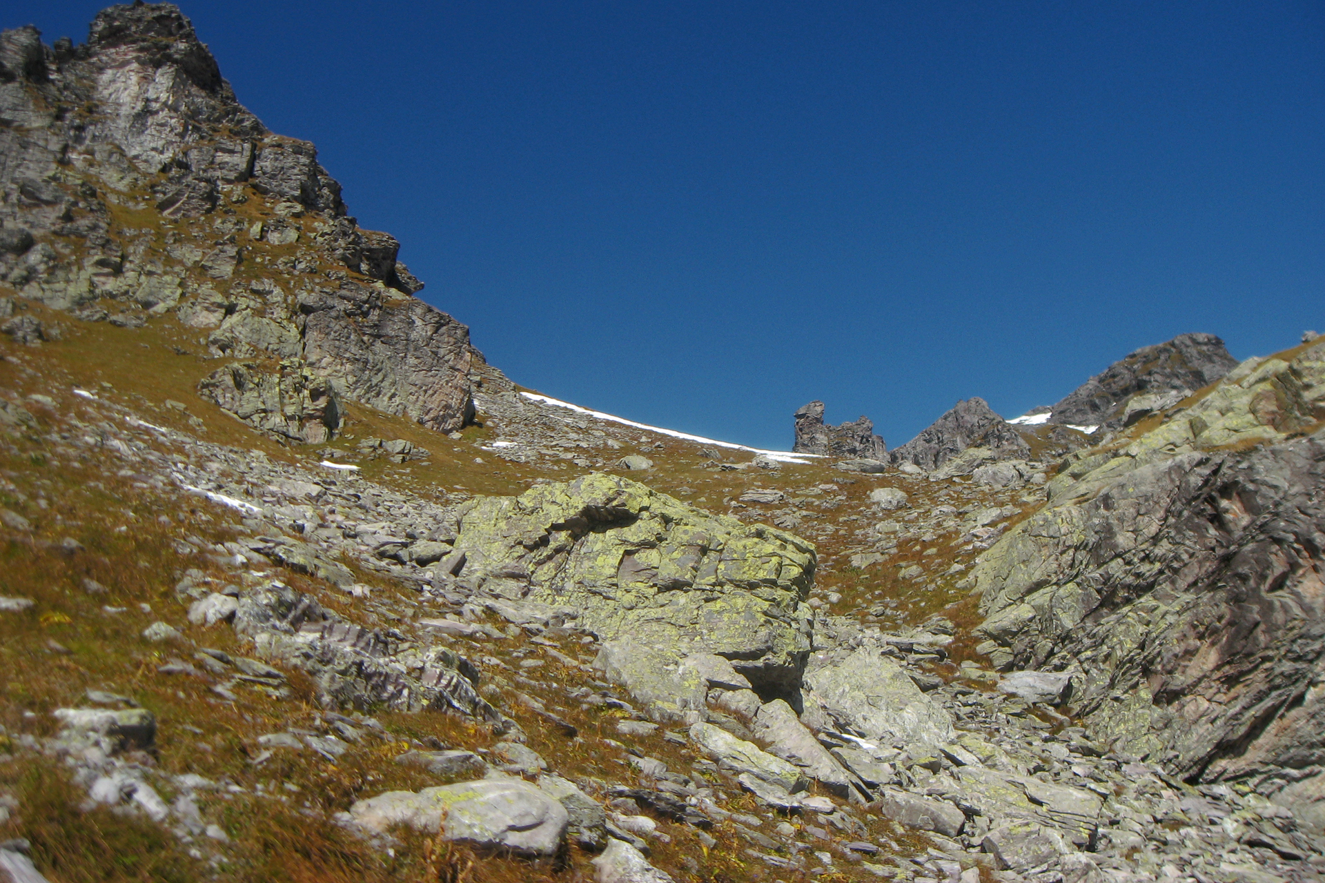 Fotografia przedstawia krajobraz skał na tle błękitnego nieba. Między szarymi skałami rośnie zrudziała trawa. Na skałach rosną żółte i szare porosty skorupiaste.