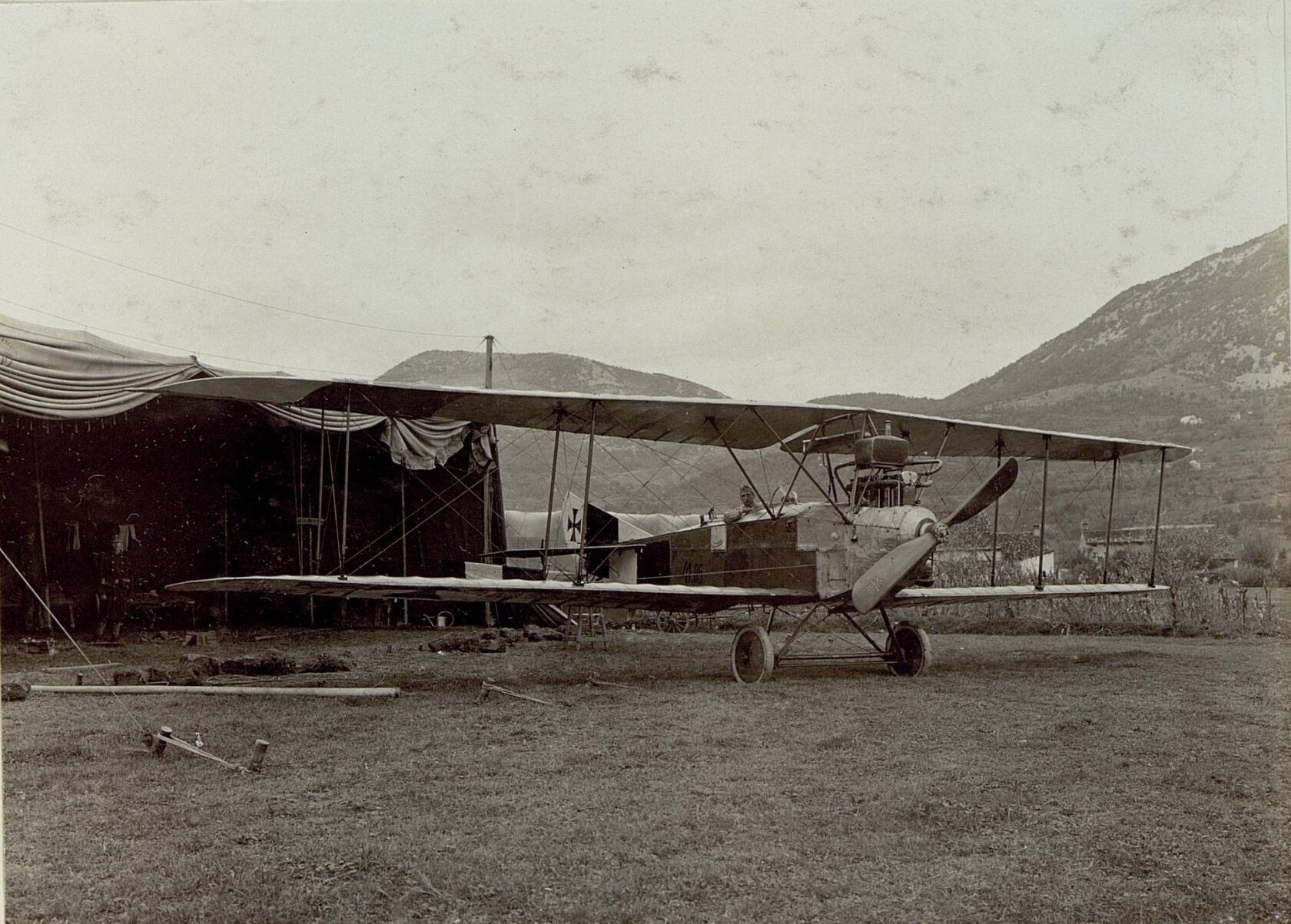 Na ilustracji samolot stojący na trawie. Z tyłu rozbity obóz, widoczne wiszące kawałki materiałów jako zasłona obozu, oraz widoczne góry.