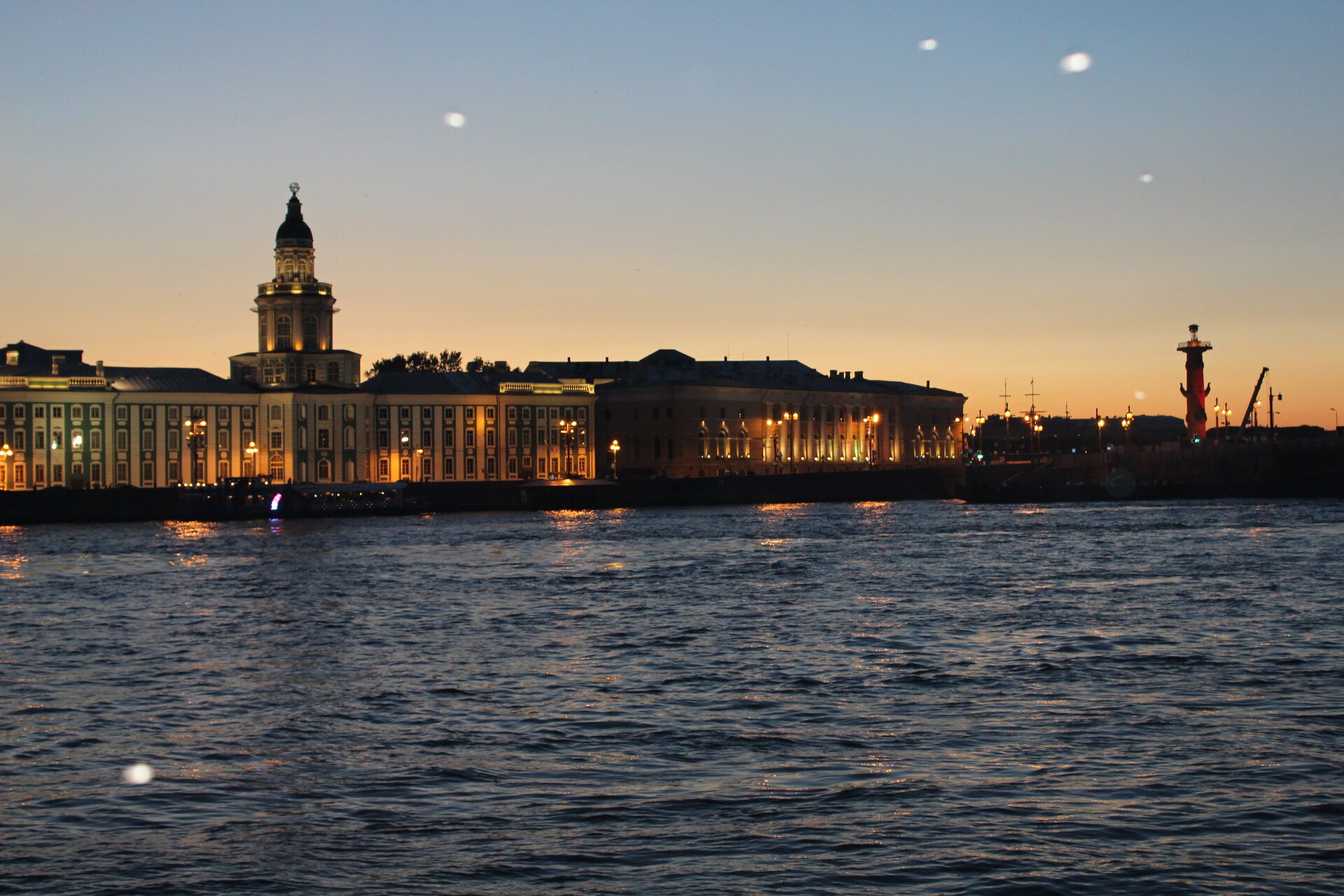 Na zdjęciu jest rzeka, na której brzegu stoi oświetlony długi budynek z wieżą. To Muzeum Ermitażu. Na zdjęciu panuje zmierzch.   