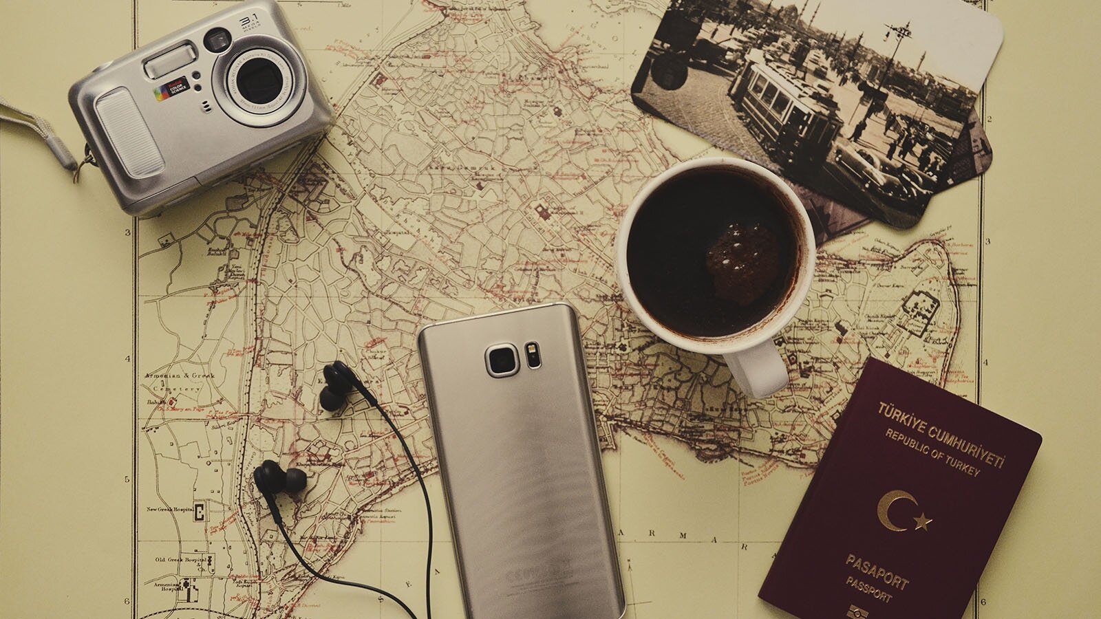 Zdjęcie przedstawia czarnobiałą mapę, na której ułożono: paszport, aparat fotograficzny, telefon, słuchawki wkładane do uszu, filiżankę czarnej kawy i zdjęcie starego tramwaju. 