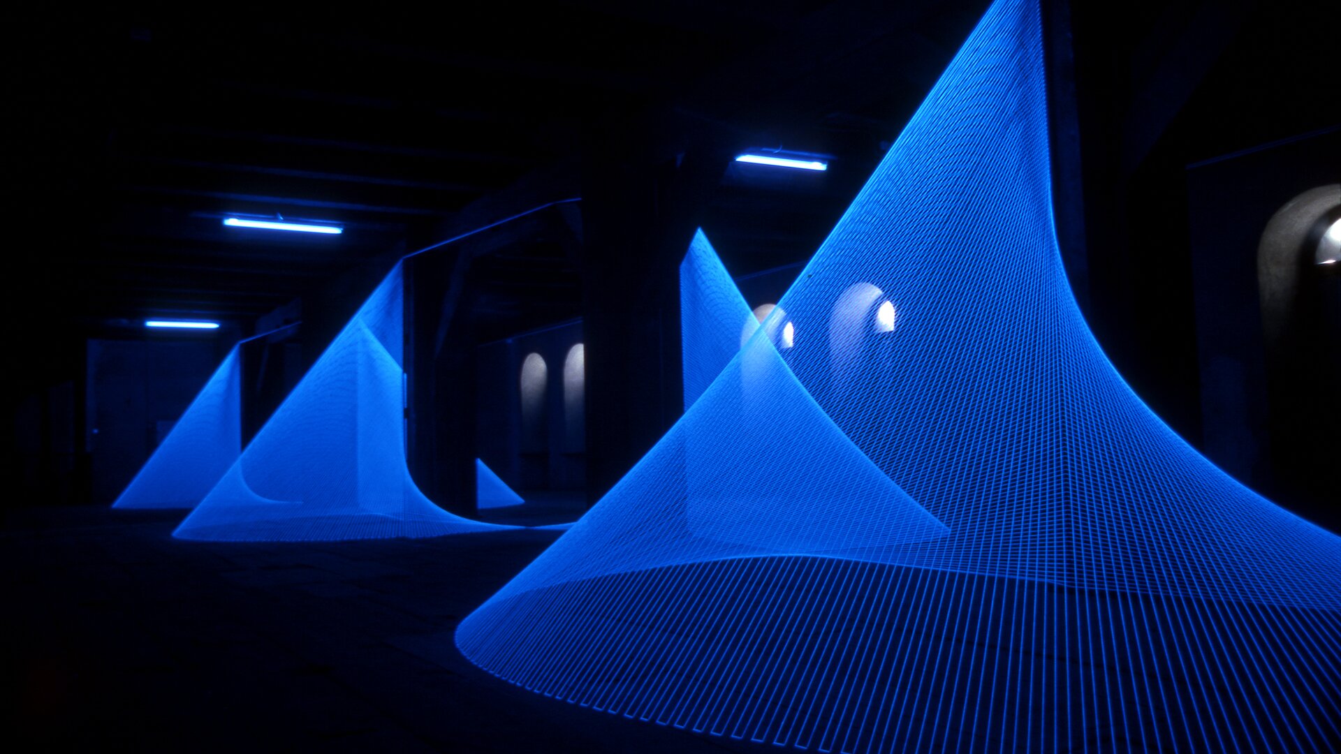 Na zdjęciu jest ukazane ciemne pomieszczenie z kilkoma instalacjami w kształcie stożka o kolorze niebieskim. Nad instalacją znajdują się 3 wąskie, podłużne lampy. W tle są łukowate okna.