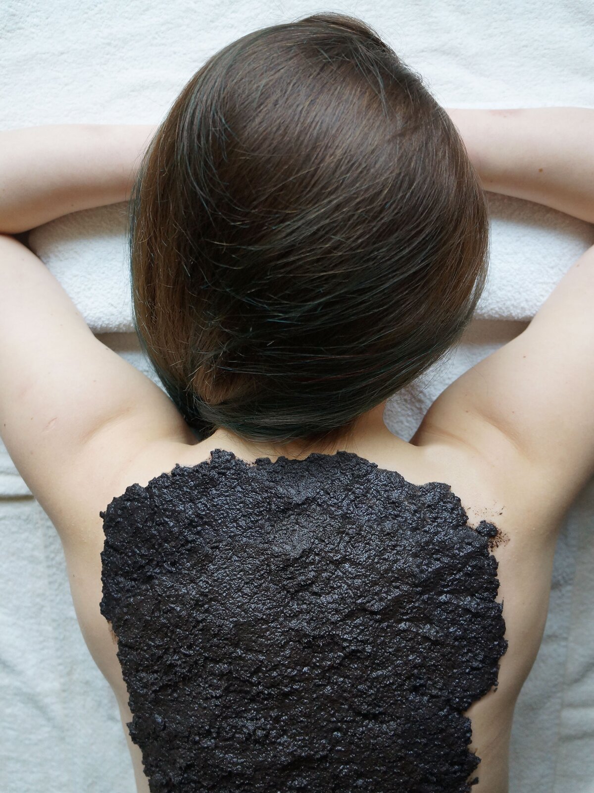 Fotografia przedstawiająca błotną masę rozsmarowaną na plecach kobiety. Jest to okład borowinowy.