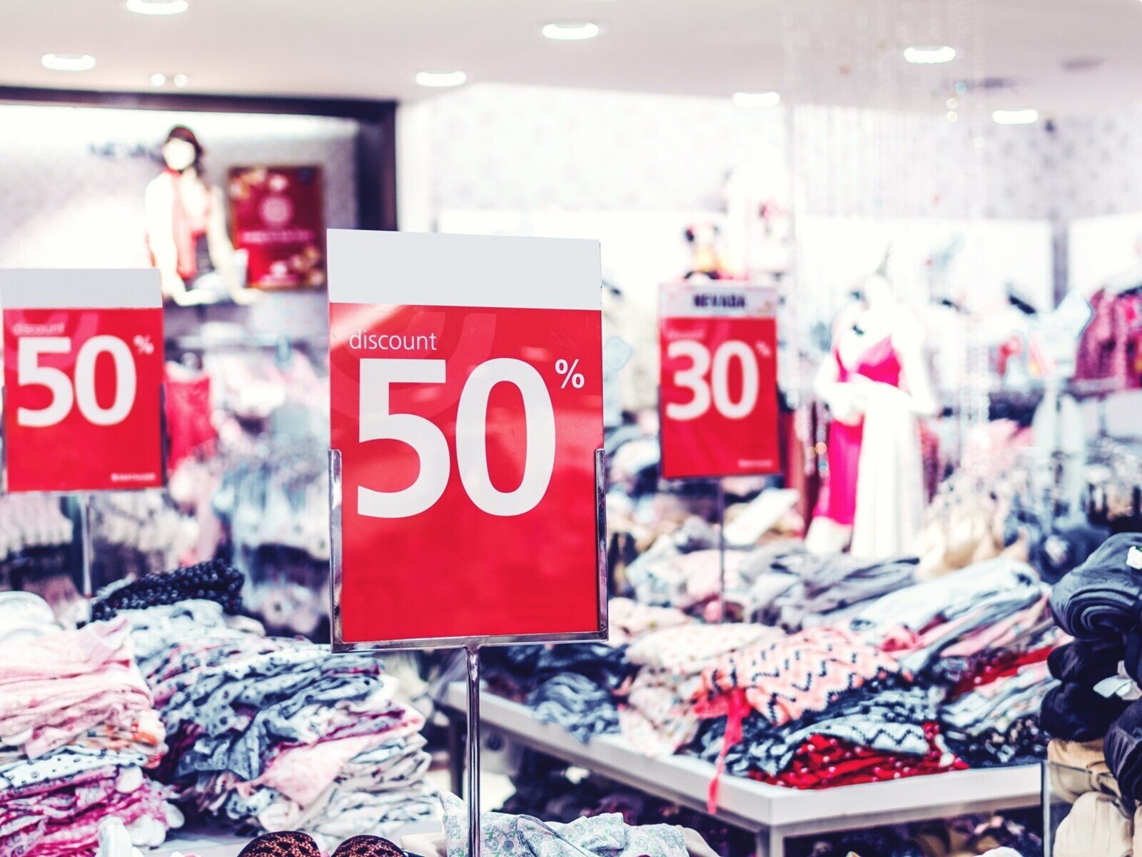 Zdjęcie przedstawia sklep z odzieżą. Widoczne są stoły, na których ułożono ubrania. Przy każdym stole ustawiono tabliczki, na których widoczne są cyfry o ile produkt został przeceniony.  Na czerwonym tle widnieje napis 50 % obok 30%  oraz napis w języku angielskim DISCOUNT. 