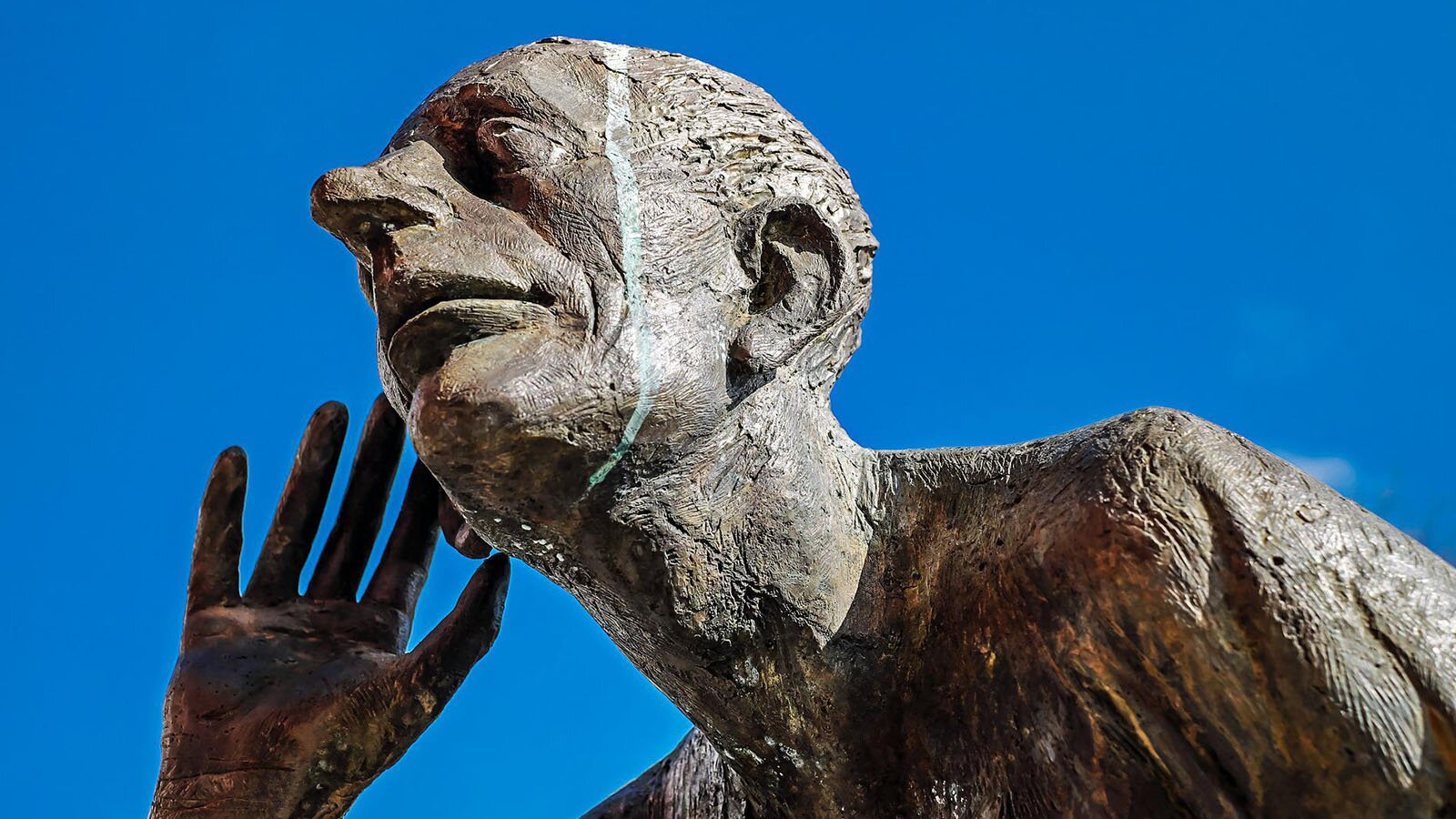 Zdjęcie przedstawia fragment rzeźby człowieka. Widoczna jest jego głowa, prawa dłoń oraz ramiona. Głowa jest uniesiona ku górze. Widoczny jest długi nos, duże oczy z opadającymi zewnętrznymi kącikami, szerokie usta. Prawa dłoń znajduje się na wysokości prawego ucha. Kciuk, palec wskazujący i środkowy dotykają ucha w geście nasłuchiwania.