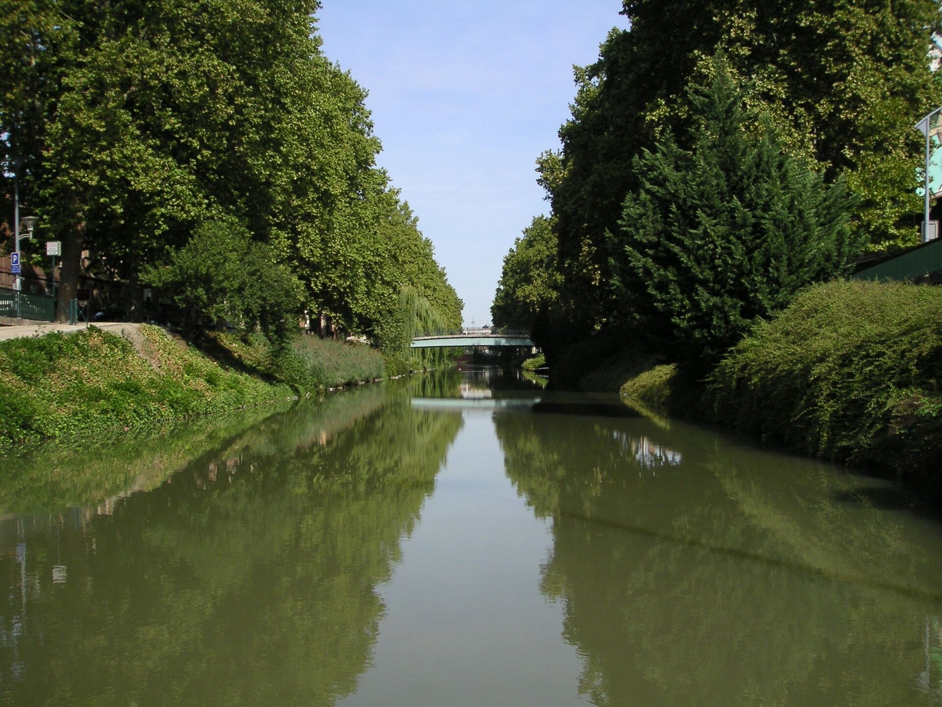 Zdjęcie przedstawia kanał. Pośrodku znajduje się płynąca woda, z dwóch stron rozpościerają się drzewa, które odbijają się w lustrze wody. Między dwoma brzegami kanału rozpościera się most.