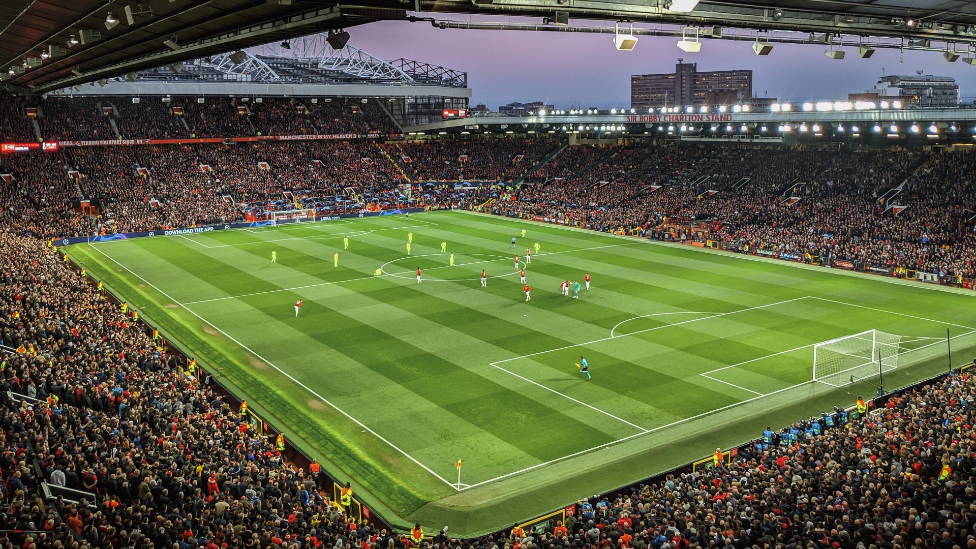 Zdjęcie przedstawia oświetlony stadion pełen kibiców w czasie meczu piłki nożnej.