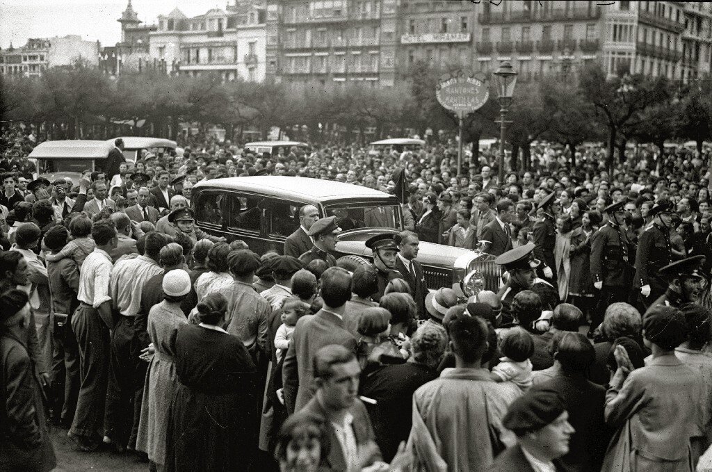 Zdjęcie przedstawia przejeżdżający samochód, który jest otoczony przez tłum ludzi. W tle znajdują się kamienice.