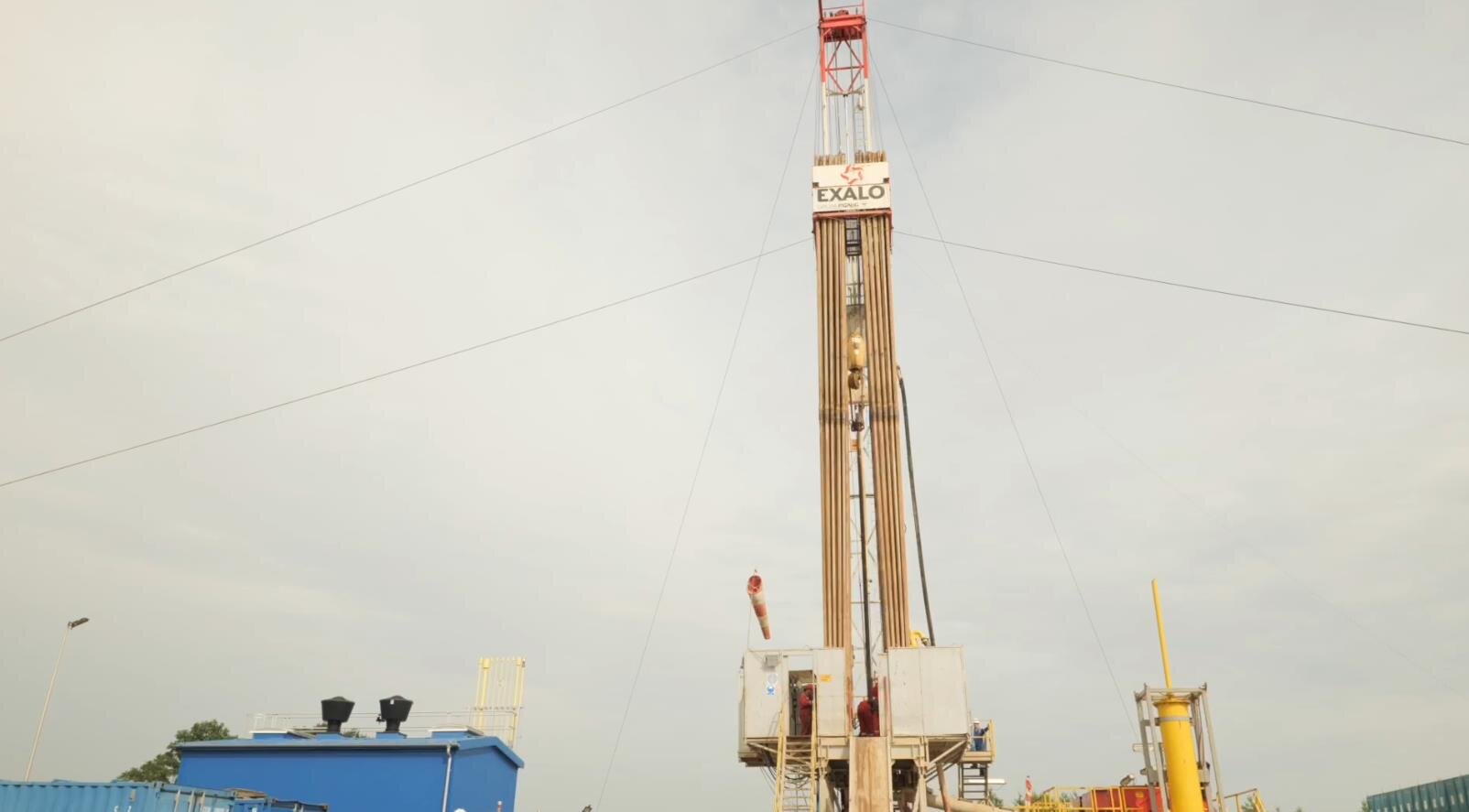 Na zdjęciu widać maszt urządzenia wiertniczego. Widoczne na zdjęciu urządzenie wiertnicze jest w kształcie ostrosłupa ściętego. W dolnej części zdjęcia widoczny jest podest na którym znajdują się dwa białe kontenery. Pomiędzy kontenerami umieszczona jest wieża wiertnicza. Wieża jest konstrukcją kratową o kształcie cienkiego podłużnego prostokąta o orientacji pionowej. Z boku wieży odchodzą stalowe liny. Są to odciągi. Po bokach wieży zamontowane są długie rury. Jest to ich miejsce magazynowe. Na górze wieży widać walec po którym biegnie w dół stalowa lina. Jest to wielokrążek górny. Na końcu liny widoczne jest urządzenie do którego przyłączona jest rura wiertnicza. 