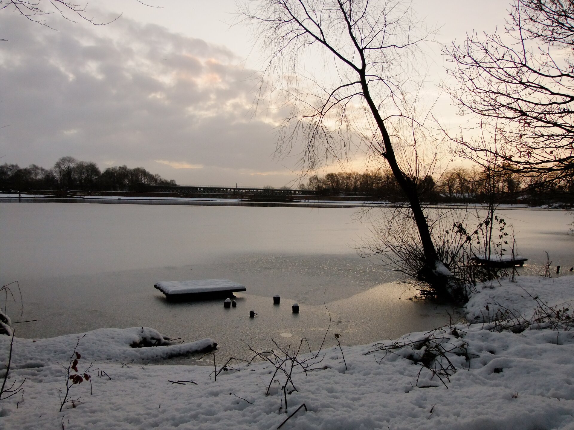 Fotografia wykonana zimą prezentuje duże jezioro oraz fragment jego brzegu okrytego śniegiem. Tafla jeziora jest pokryta lodem, na którym leży śnieg. Przy brzegu lód wydaje się cienki. W oddali widać niezamarznięte jeszcze wody jeziora.