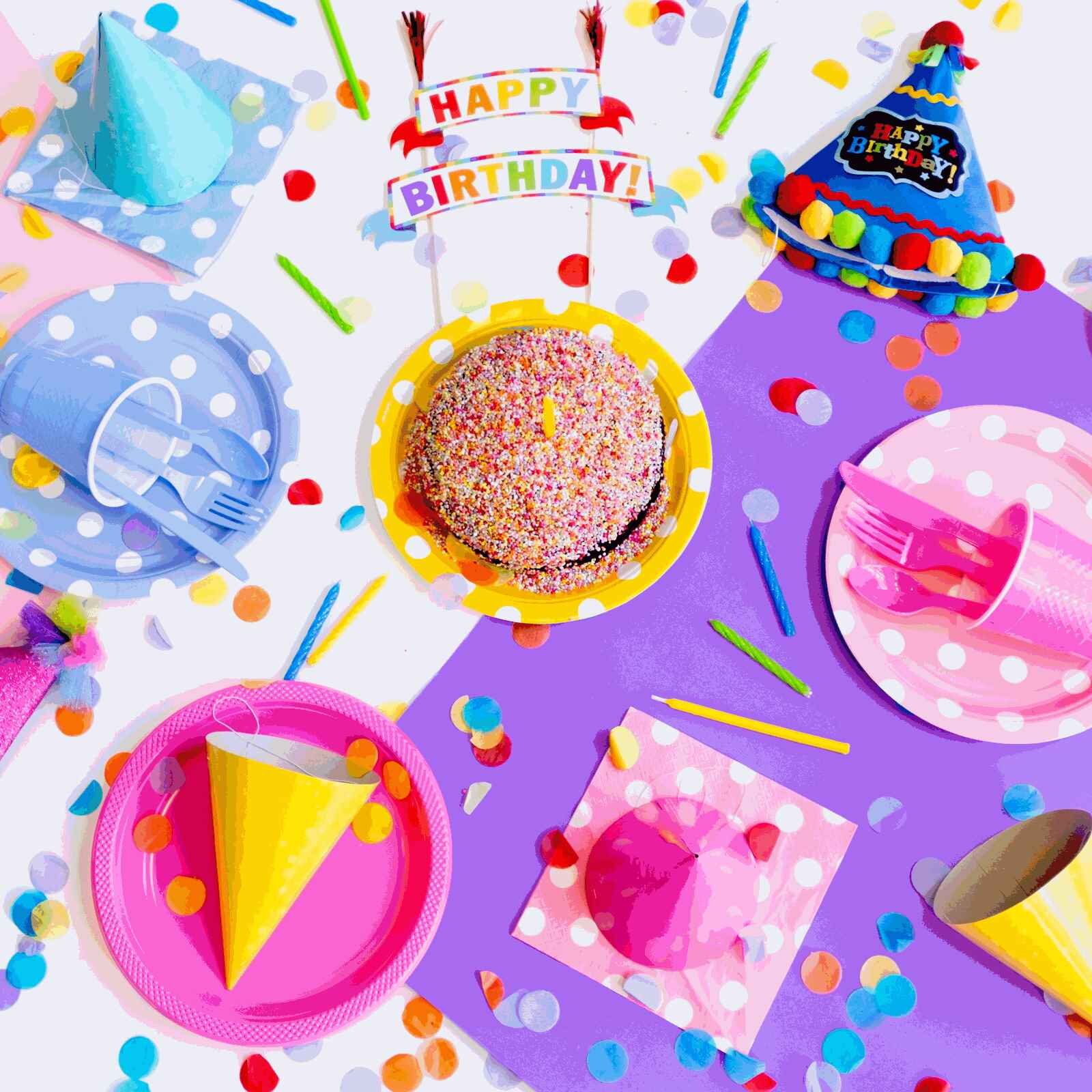 Zdjęcie przedstawia stół na środku którego jest tort posypany kolorową posypką, obok leżą czapeczki urodzinowe i papierowe talerze i sztućce, nad tortem jest napis "HAPPY BIRTHDAY".