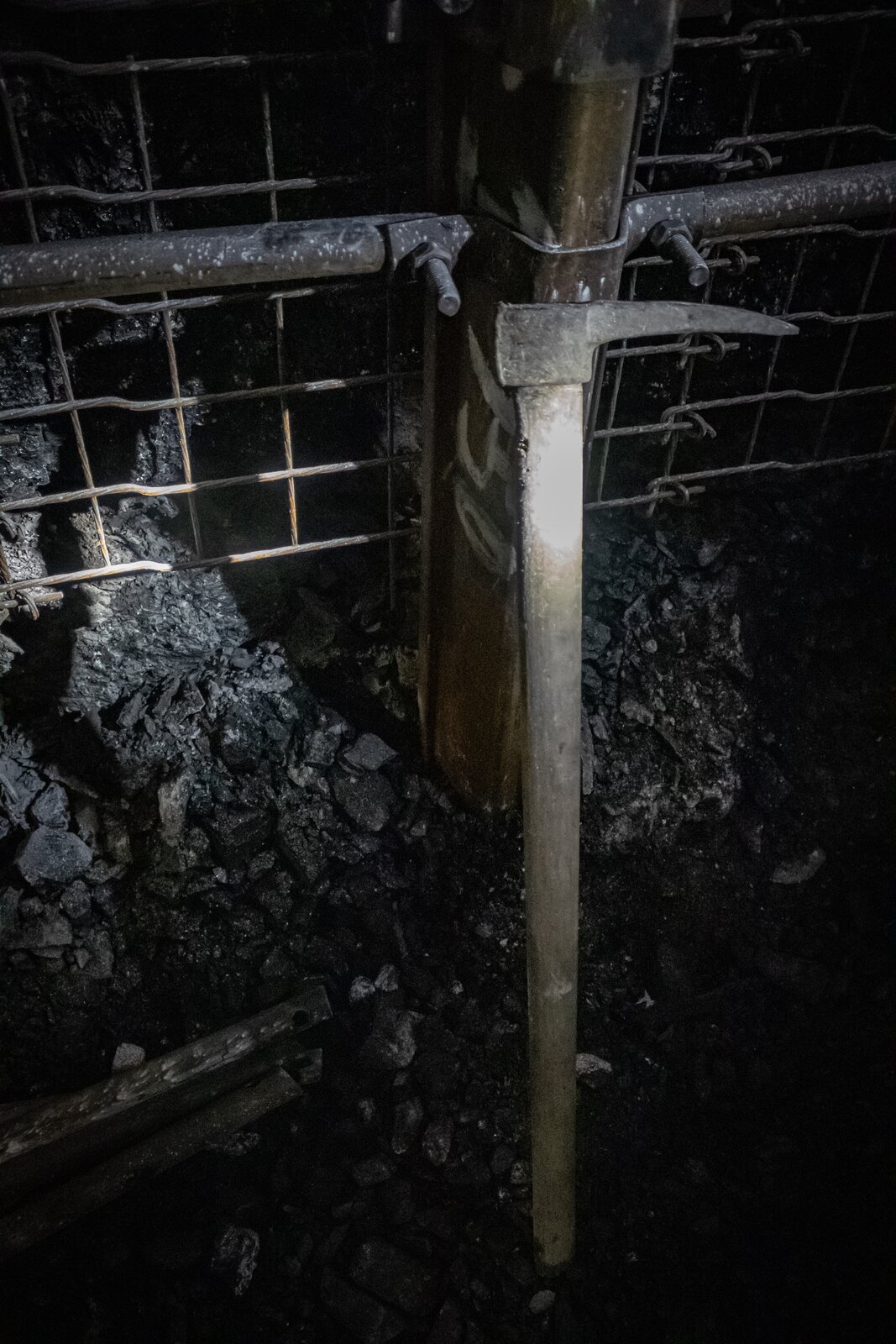 Na zdjęciu widać kilof górniczy. Kilof jest o kształcie zbliżonym do litery L. Trzon kilofa jest drewniane. Organ roboczy stalowy. Organ jest krótki. Kilof oparty jest o siatkę zabezpieczającą ocios.