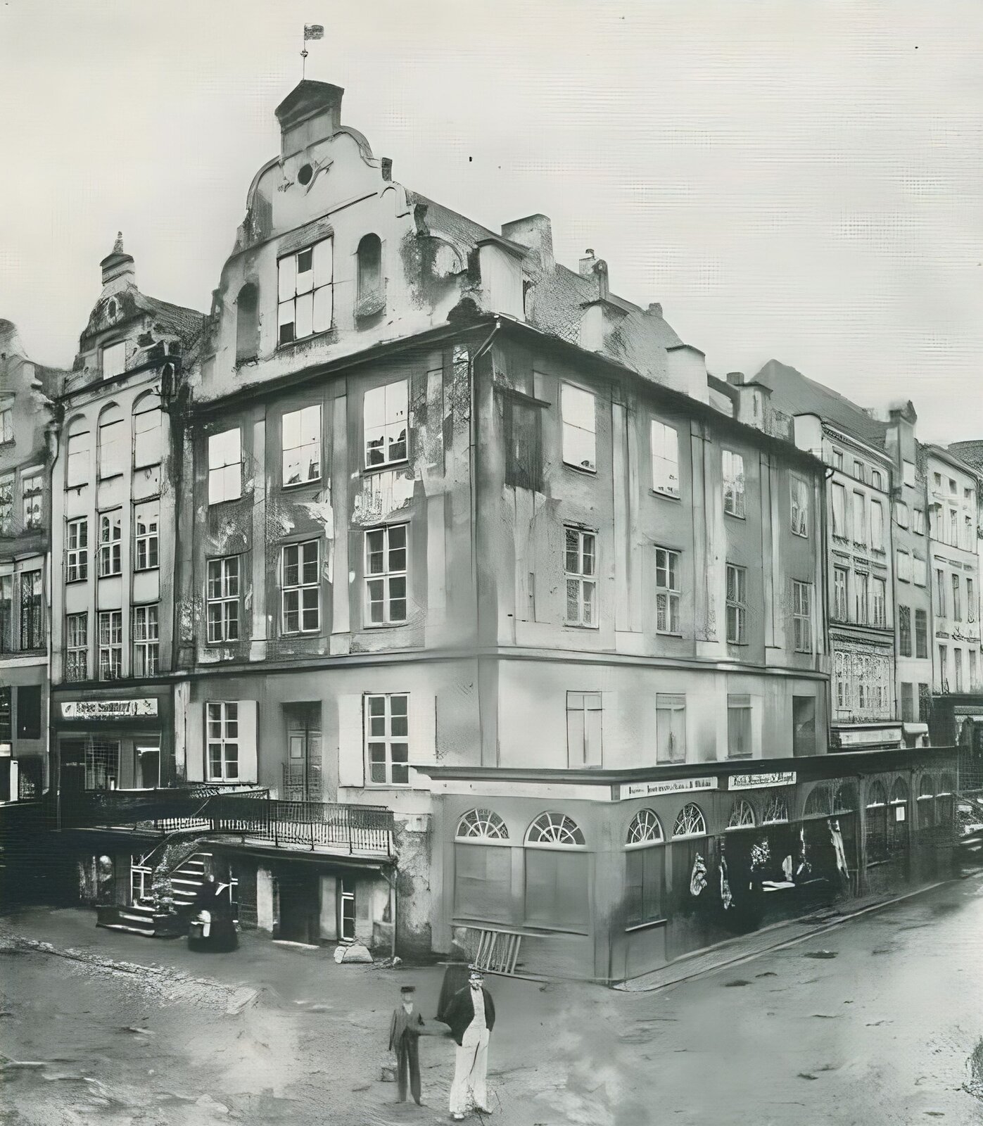 Ilustracja przedstawia wykonane w XIX wieku zdjęcie elbląskiego Dworu Artusa - kamienicy narożnej z przedprożem. Pod przedprożem kamienicy wejście do oficyny budynku. Prostokątne okna flankują drzwi wejściowe ulokowane na wysokim parterze. W górnych kondygnacjach dwa rzędy okien. Szczyt budynku utrzymany w stylu manieryzmu niderlandzkiego - ornament okuciowy na skraju konstrukcji szczytu oraz niewielka lukarna. Przy ścianie bocznej w parterze, na całej długości znajduje się konstrukcja drewnianej oficyny z afiszami, okna prostokątne są zasłonięte, nad każdym oknem świetlik w formie połowy rozety. U dołu widać grupę przechodniów. Od lewej i prawej strony fotografii widoczne kolejne pierzeje kamienic. Droga pokryta kostką brukową.