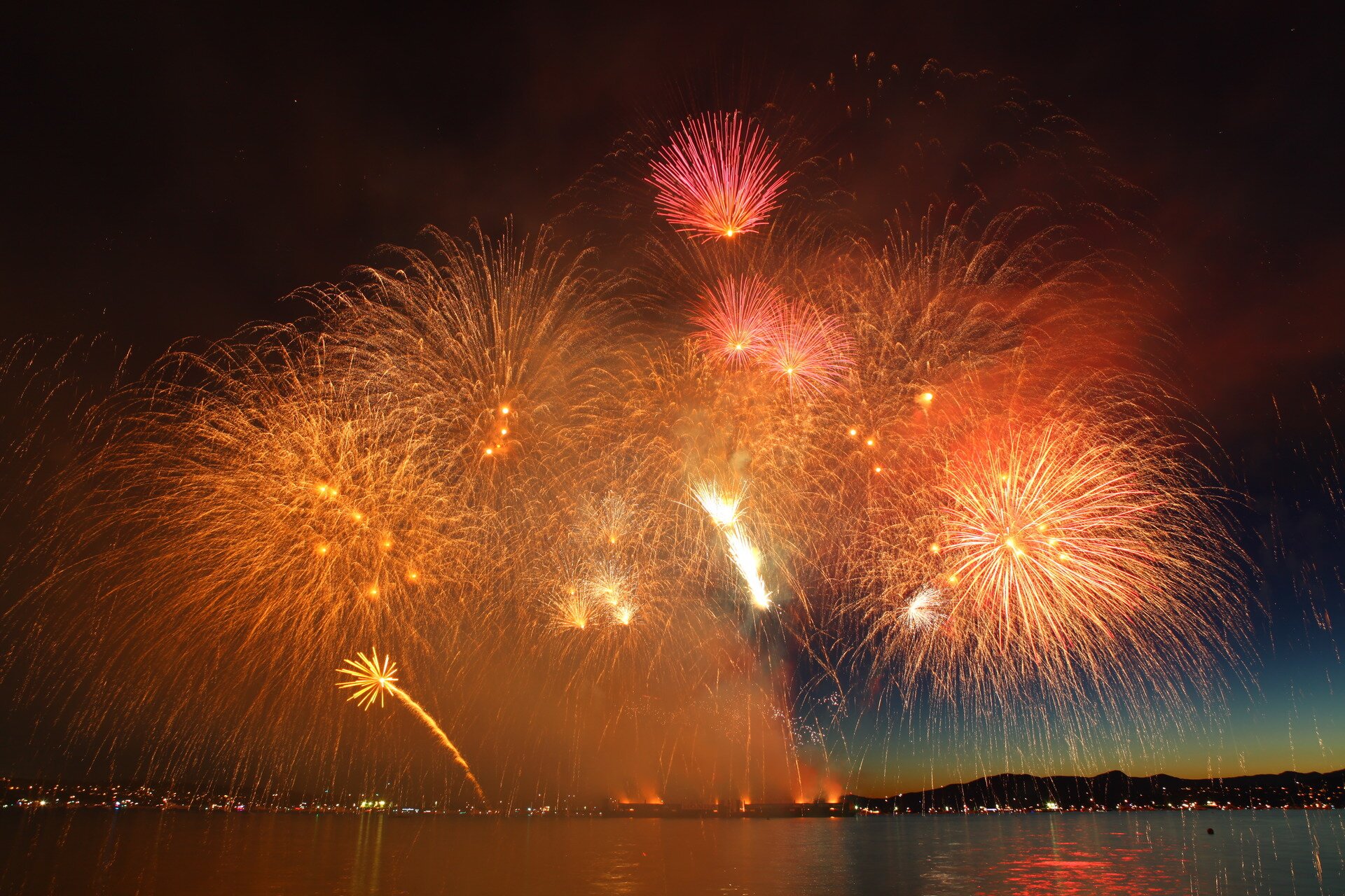 Zdjęcie przedstawia nocne niebo nad miastem leżącym nad jeziorem lub brzegiem morza i bardzo efektowny pokaz fajerwerków.
