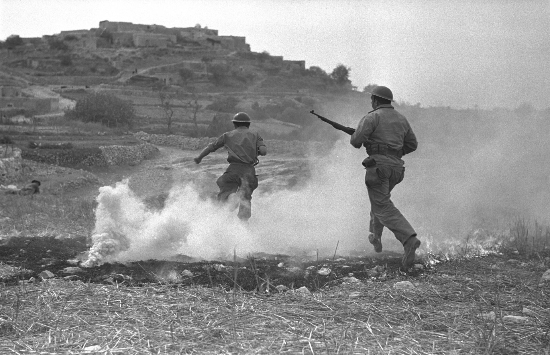 Ilustracja przedstawia dwóch biegnących mężczyzn w hełmach oraz strojach wojskowych. Jeden z nich trzyma karabin. Obok nich na ziemi leży granat, z którego wydobywa się jasny dym. W tle znajdują się budynki skały i droga. Zdjęcie jest czarno-białe.
