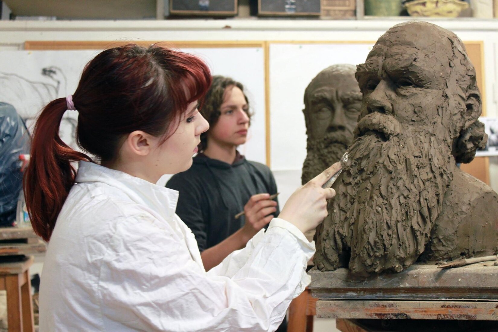 Fotografia przedstawia postaci pracujące przy rzeźbach. Są to kobieta oraz mężczyzna. Kobieta widoczna jest na pierwszym planie z profilu, ma na sobie białą, wygniecioną koszulę, włosy związane w kucyk oraz spojrzenie skierowane ku rzeźbie. Kobieta stoi z lewej strony, rzeźba z prawej. Jej praca to popiersie brodatego starszego mężczyzny, wykonane z gliny. Za kobietą uchwycono mężczyznę o kręconych włosach. Ma on na sobie bluzę z kapturem, w dłoni trzyma szpatułkę, którą poprawia detale. Patrzy na podobną rzeźbę, widoczną na stanowisku obok. Tło stanowi pomieszczenie.
