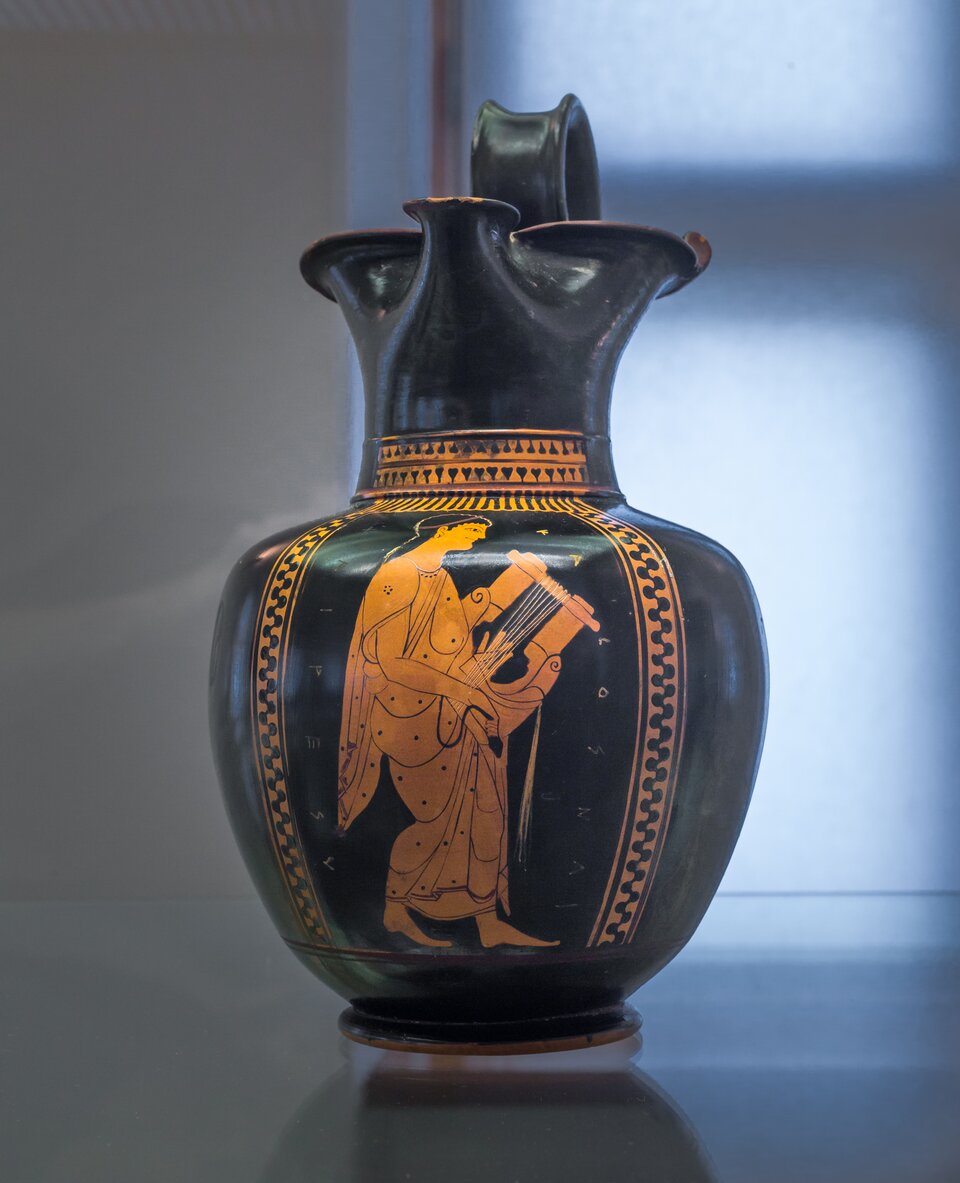 Ilustracja przedstawia czerwonofigurową wazę. W centrum dekoracji znajduje się sylwetka młodego mężczyzny grającego na kitarze. Mężczyzna stoi, trzyma instrument w rękach. Postać ubrana jest w długą szatę, jest skupiona na muzyce.